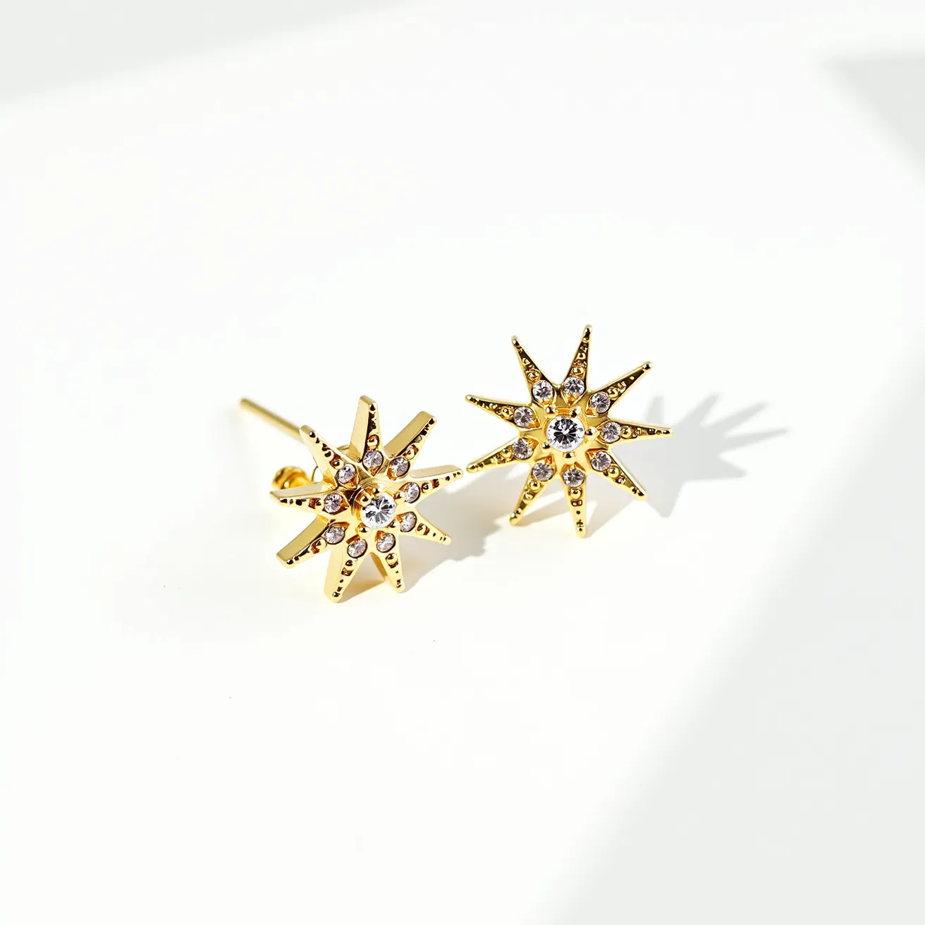 These star stud earrings are crafted from a bright gold metal, featuring an intricate design of radiating star points. Each earring is adorned with small, clear gemstones, possibly cubic zirconia, placed meticulously at the tip of each point and surrounding a larger central stone. The stones are round cut, providing a brilliant sparkle that complements the golden hue of the metal. The gemstones are set in a secure prong setting, ensuring they remain firmly in place while adding elegance to the overall design. The earrings are equipped with a simple post back, suitable for pierced ears, offering a classic and secure attachment style.