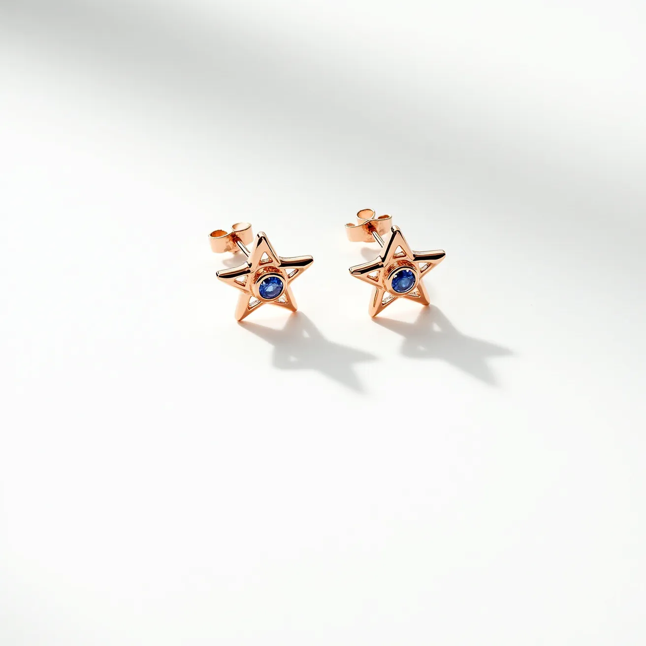 These star stud earrings are crafted with a rose gold metal that forms the shape of a five-pointed star. In the center of each star, there is a round-cut blue gemstone, possibly a sapphire, securely set in a bezel setting. The earrings have a post and butterfly clasp attachment, ensuring a secure fit when worn.