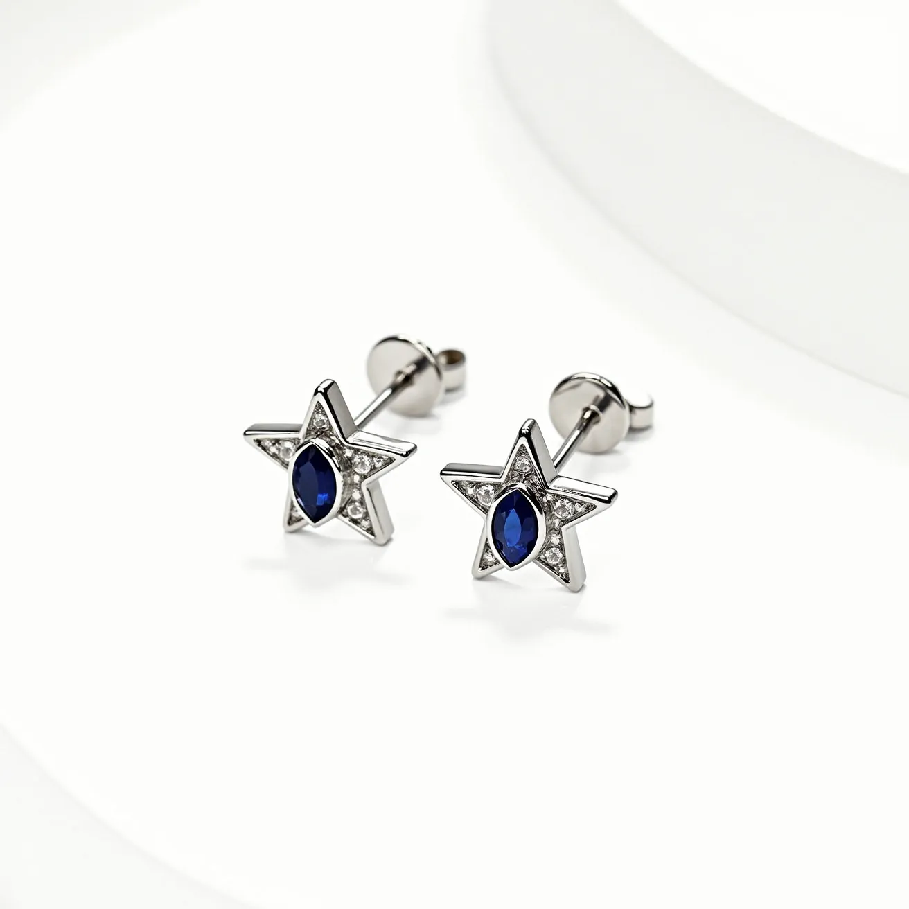 These star stud earrings feature a sleek metallic design with each earring shaped into a five-pointed star. At the center of each star is a deep blue, marquise-cut gem, set with precision. Surrounding the central gem are several smaller round clear stones, creating a striking contrast and adding extra sparkle to the piece. The earrings use a classic push-back clasp, ensuring secure and comfortable wear. The combination of the polished metal, vivid center gem, and the surrounding stones gives these earrings an elegant and contemporary appeal.