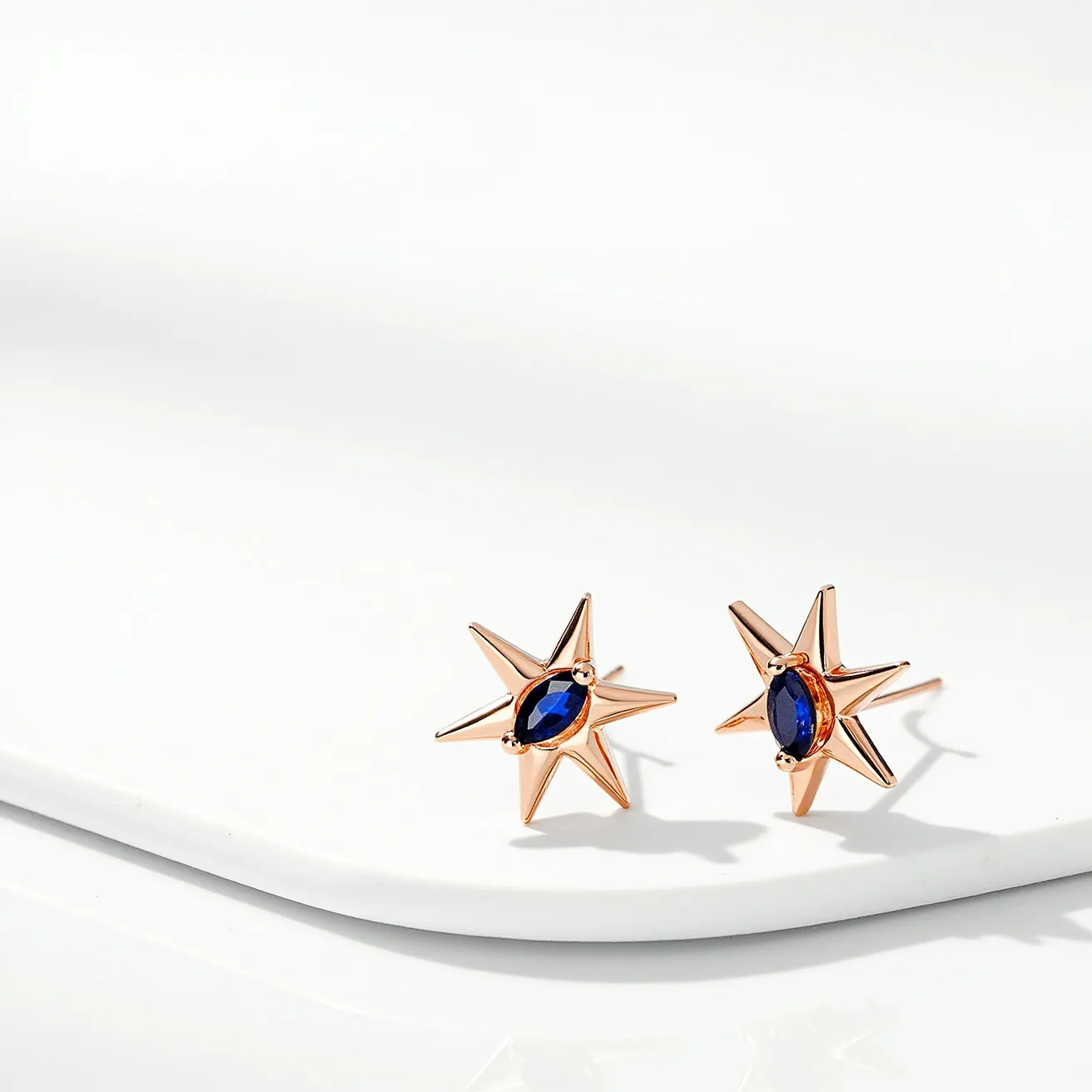 These star stud earrings are elegantly crafted with a brilliant rose gold metal, forming a striking star design. Each earring features a deep blue, marquise-cut gemstone, expertly set at the center of the star, adding an element of sophistication and depth. The stones are held securely in place with a bezel or similar setting, ensuring they remain the focal point. The earrings are designed to be worn with a simple and classic post stud attachment, providing ease and comfort for the wearer.