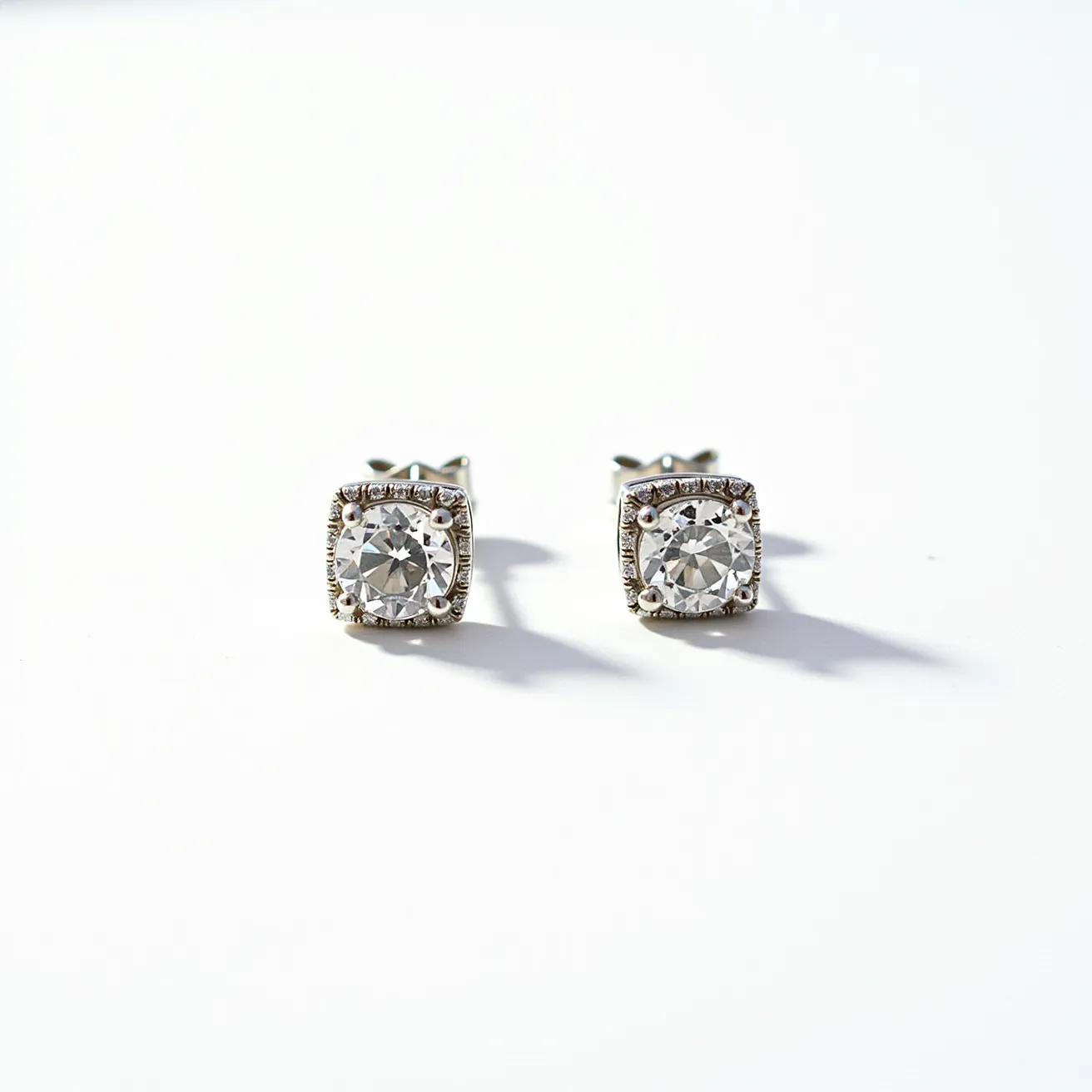 These stud earrings feature a central round-cut gemstone, which appears to be a sparkling diamond or diamond simulant. The stones are held in place by four prongs, enhancing their brilliance and visibility. Surrounding the central stones is a halo of smaller gems set in a square formation, providing an elegant and classic frame that contrasts with the round center stones. The surrounding stones enhance the overall sparkle and give the earrings a sophisticated touch. The metal used for the setting and posts appears to be shiny, possibly silver or white gold, adding to the luxurious appearance. The earrings are attached with a butterfly (or friction) back, ensuring they stay securely in place when worn.