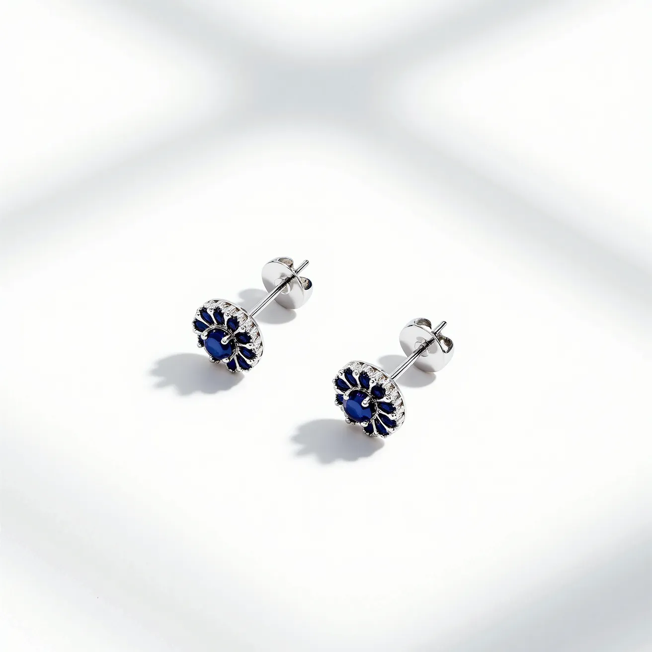 These stud earrings feature a vibrant blue gemstone, likely a sapphire, as their focal point, set in a round cut. The stones are encircled by a decorative metal setting that may incorporate small clear gems or detailing, adding a touch of brilliance and contrast to the deep blue center. The earrings are crafted from a shiny metal, possibly white gold or platinum, providing a sleek and elegant finish. They are equipped with a classic post and butterfly back clasp, ensuring secure and comfortable attachment when worn.