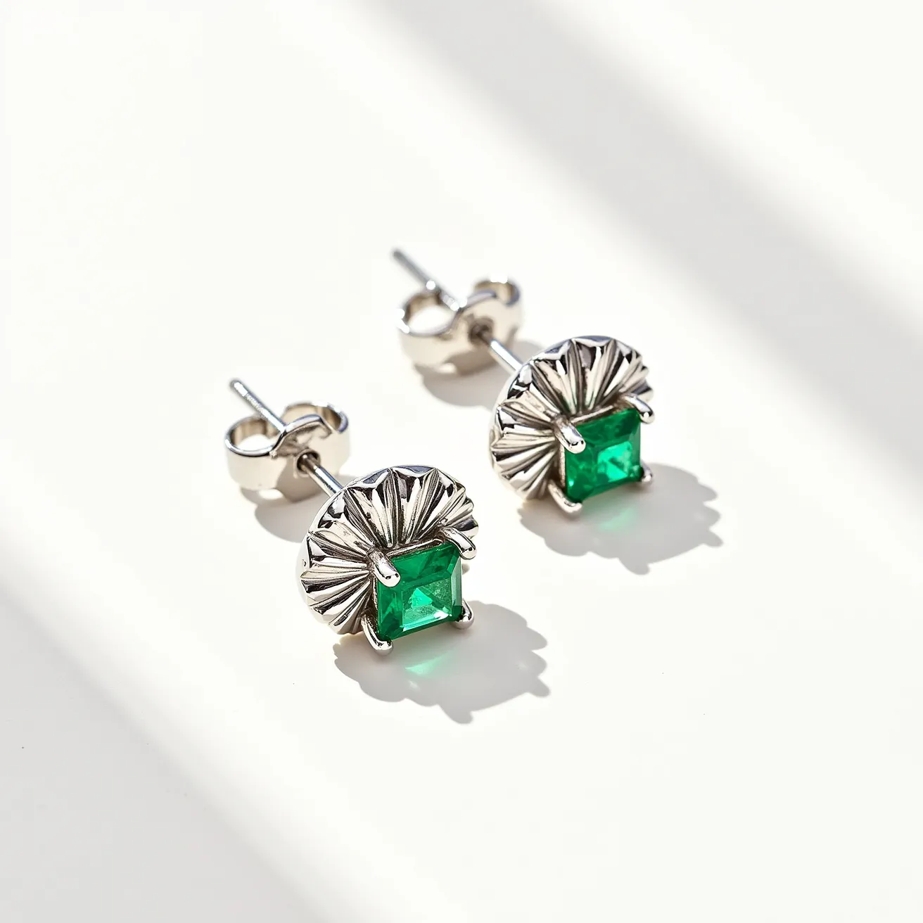 These stud earrings feature a striking green gemstone with an emerald cut, set in a unique decorative mount. The gem is securely held in place by four prongs, and the setting appears to be crafted from a silvery metal, likely sterling silver or white gold, which enhances the stone's vibrant color. The design incorporates a fan-like detail surrounding the gemstone, adding a touch of elegance and texture to the studs. They employ a standard post back with butterfly clasps for secure and comfortable wear, making them a stylish choice for everyday or special occasions.