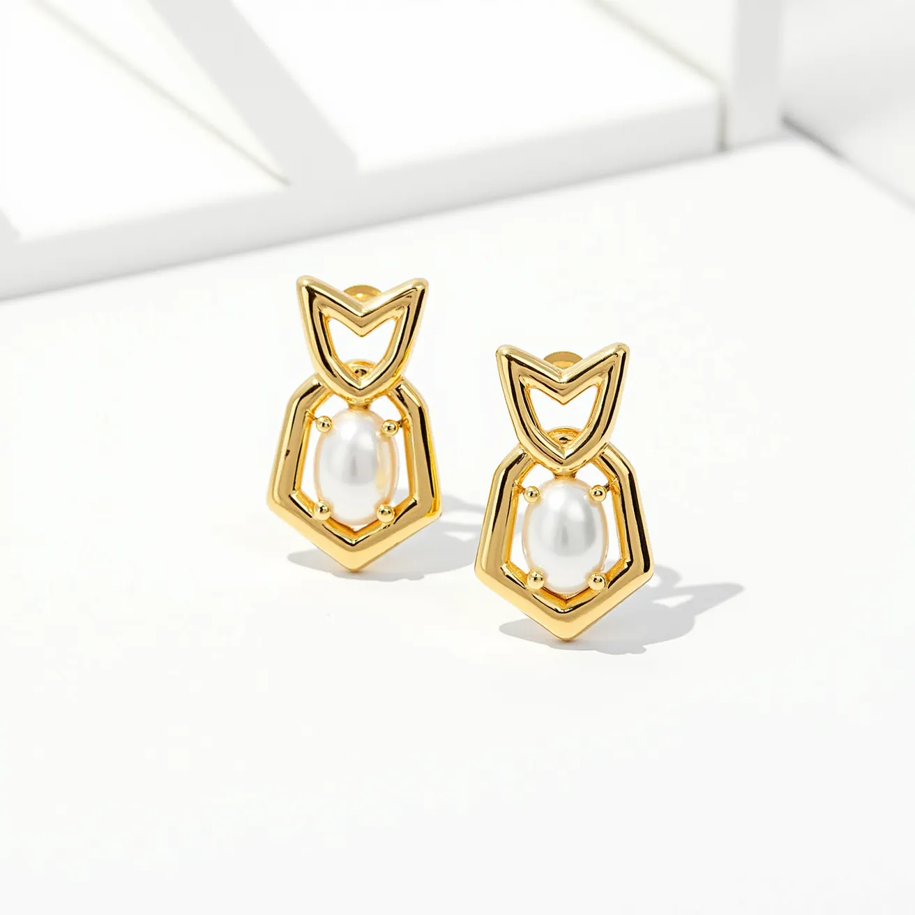 These stud earrings feature a geometric design crafted from a glossy gold-colored material that frames a central pearl-like gem. The gem is oval-shaped and is secured within the frame by four prongs, enhancing its elegant appearance. The overall styling of the earrings includes a unique angular pattern that resembles a modern, artistic motif. They are designed to be worn with a post and butterfly clutch attachment for a secure fit. The smooth and sleek finish, combined with the lustrous gem, gives these earrings a sophisticated and contemporary appeal.