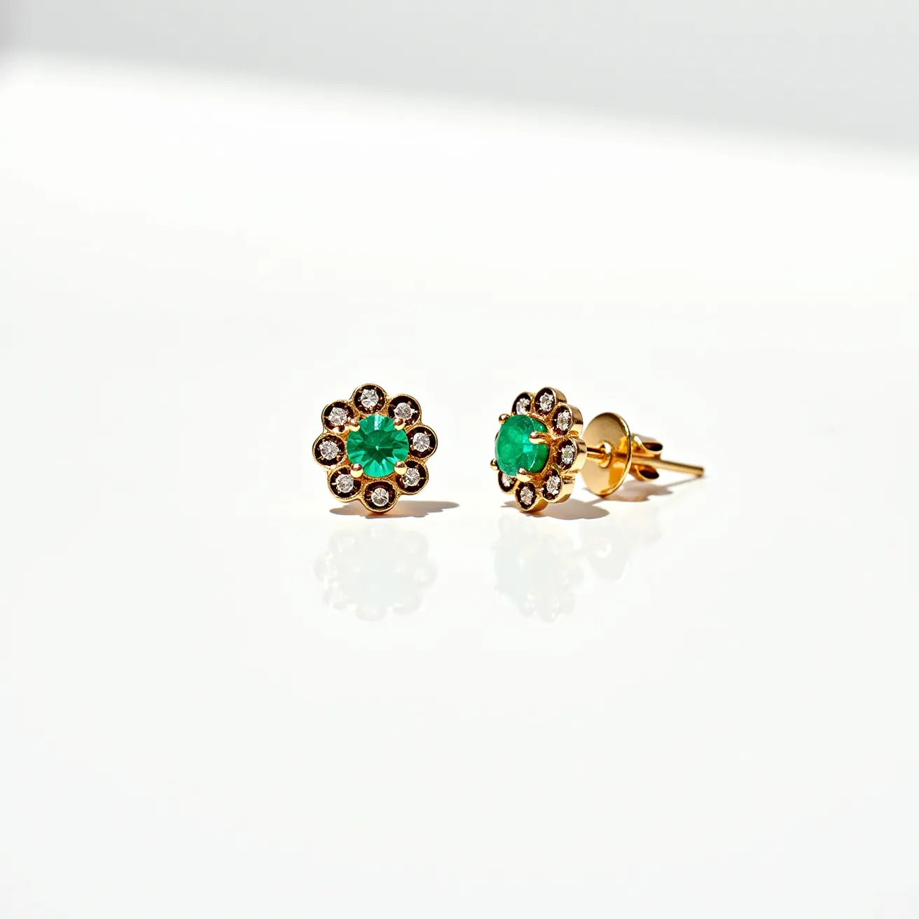 These stud earrings feature a central emerald gemstone, round in shape and prong-set, surrounded by a halo of smaller round-cut diamonds. The diamonds are also prong-set, enhancing the overall sparkle and creating a floral motif. The setting appears to be made of gold, which complements the green hue of the emerald and the brilliance of the diamonds. The studs are secured with a traditional post and butterfly clasp, ensuring they stay in place while worn. Overall, the combination of emerald and diamonds set in gold lends a timeless elegance to these earrings.