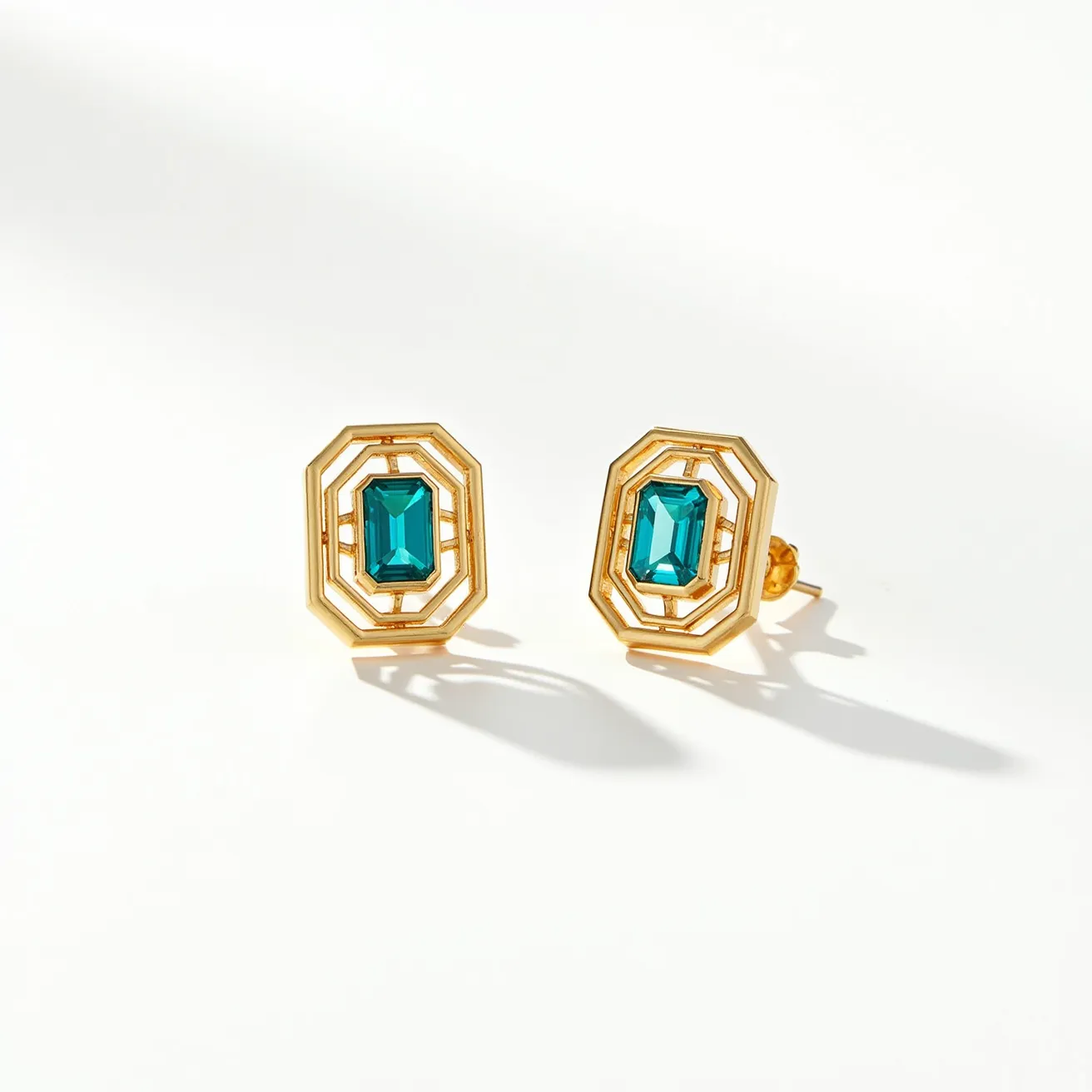 These teal earrings feature an elegant design with a prominent emerald-cut teal gemstone at the center. The stone is set within a geometric, gold-toned frame, which enhances its rich color and brilliance. The frame consists of multiple layers that create a sophisticated, openwork pattern, adding depth and interest to the design. These earrings are equipped with a post and butterfly back clasp, ensuring a secure and comfortable fit. The combination of the vibrant teal gem and the warm golden setting creates a harmonious and stylish aesthetic.