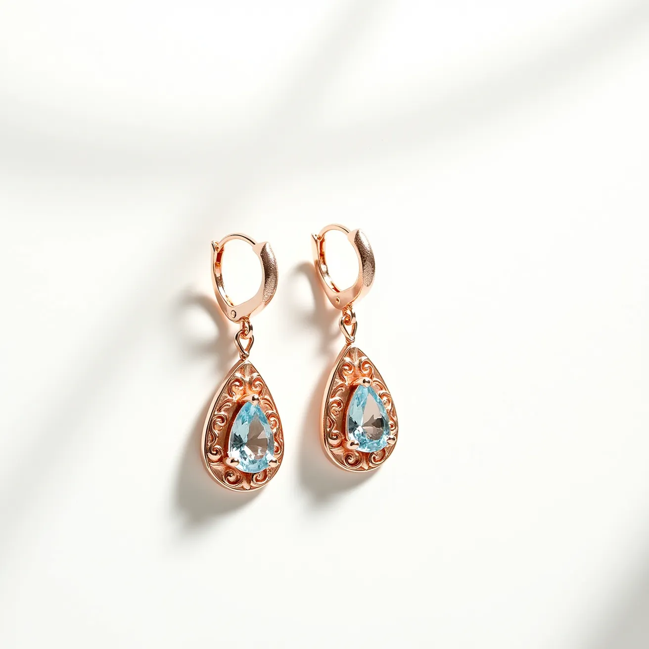 These teardrop earrings feature a delicate design with a central blue gemstone in each piece, cut in a teardrop shape. The stones are securely set within a detailed metal border with intricate scrollwork, which appears to be crafted from a warm-toned metal, possibly rose gold. The earrings are attached with hinged hoop clasps that ensure a secure fit.