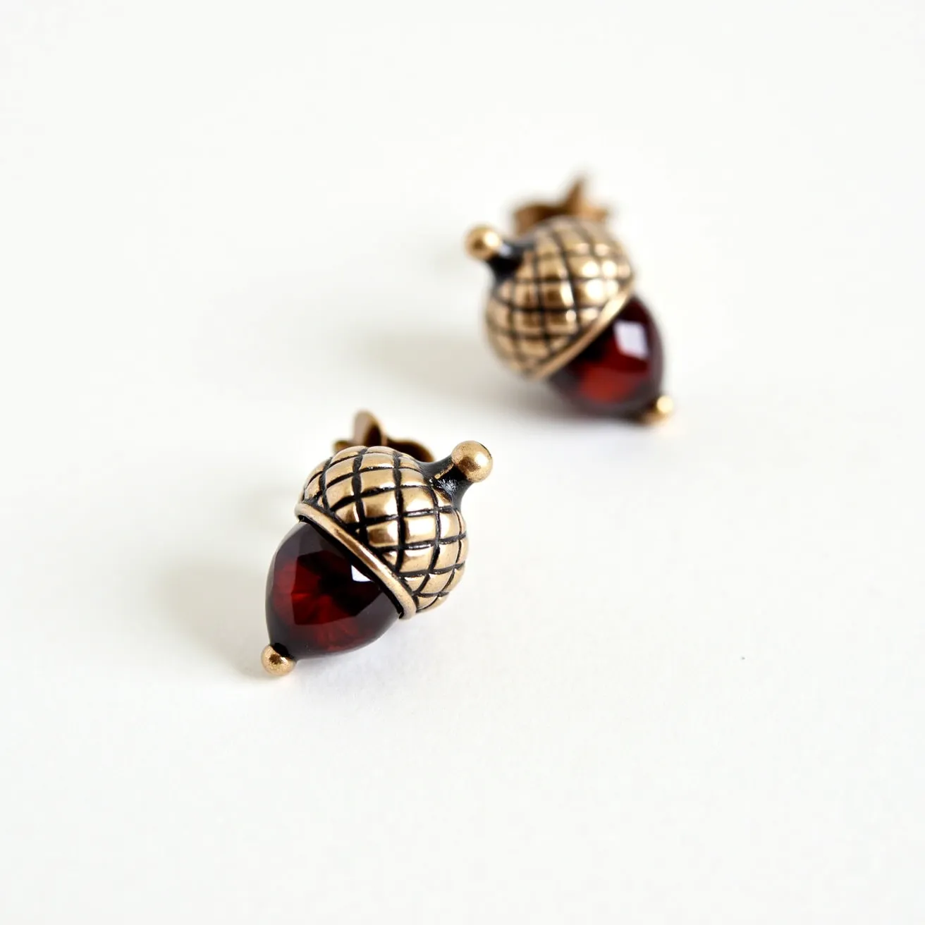 These Thanksgiving earrings feature an artistic acorn design crafted from metal with a warm, bronze-like finish. They showcase a teardrop-shaped red gemstone, likely glass or a synthetic material, with a smooth, polished surface and a deep, rich hue. The acorn cap is textured with a grid pattern, adding to the autumnal aesthetic. Each earring is designed to attach with a simple stud post and butterfly clasp, ensuring a secure fit while worn. The combination of the metallic finish and vibrant stone creates a festive and nature-inspired accessory.