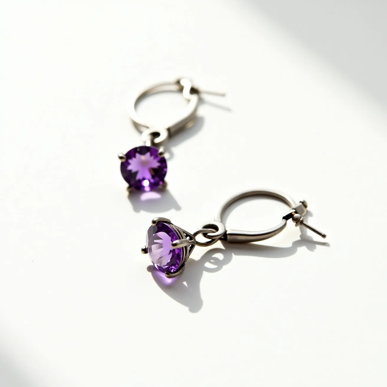 These titanium earrings feature a circular hoop design with a bright, faceted purple gemstone suspended from each hoop. The gemstones appear to be cut in a classic round shape, enhancing their sparkle and color. They are securely held in place by a prong setting, allowing maximum visibility and light reflection. The hoops are equipped with a latch back closure, ensuring both ease of use and secure wear. The overall design combines the durability of titanium with the vibrant elegance of the gemstones, resulting in a sophisticated and stylish accessory.