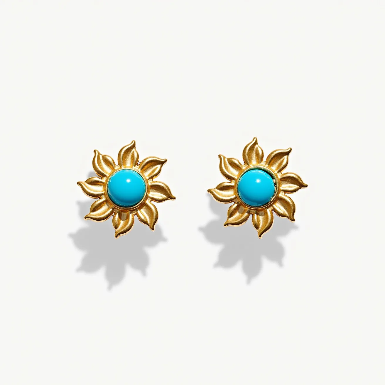 These turquoise stud earrings feature a central turquoise gemstone, round in shape, which is prominently placed in the design. The turquoise stones are set within a golden metal structure that resembles a sunburst pattern, with petal-like extensions radiating outward. The setting enhances the vibrant blue of the stones, providing a striking contrast with the warm, golden hue of the metal. The earrings likely use a typical stud attachment, securing them comfortably and stylishly on the wearer’s ears.