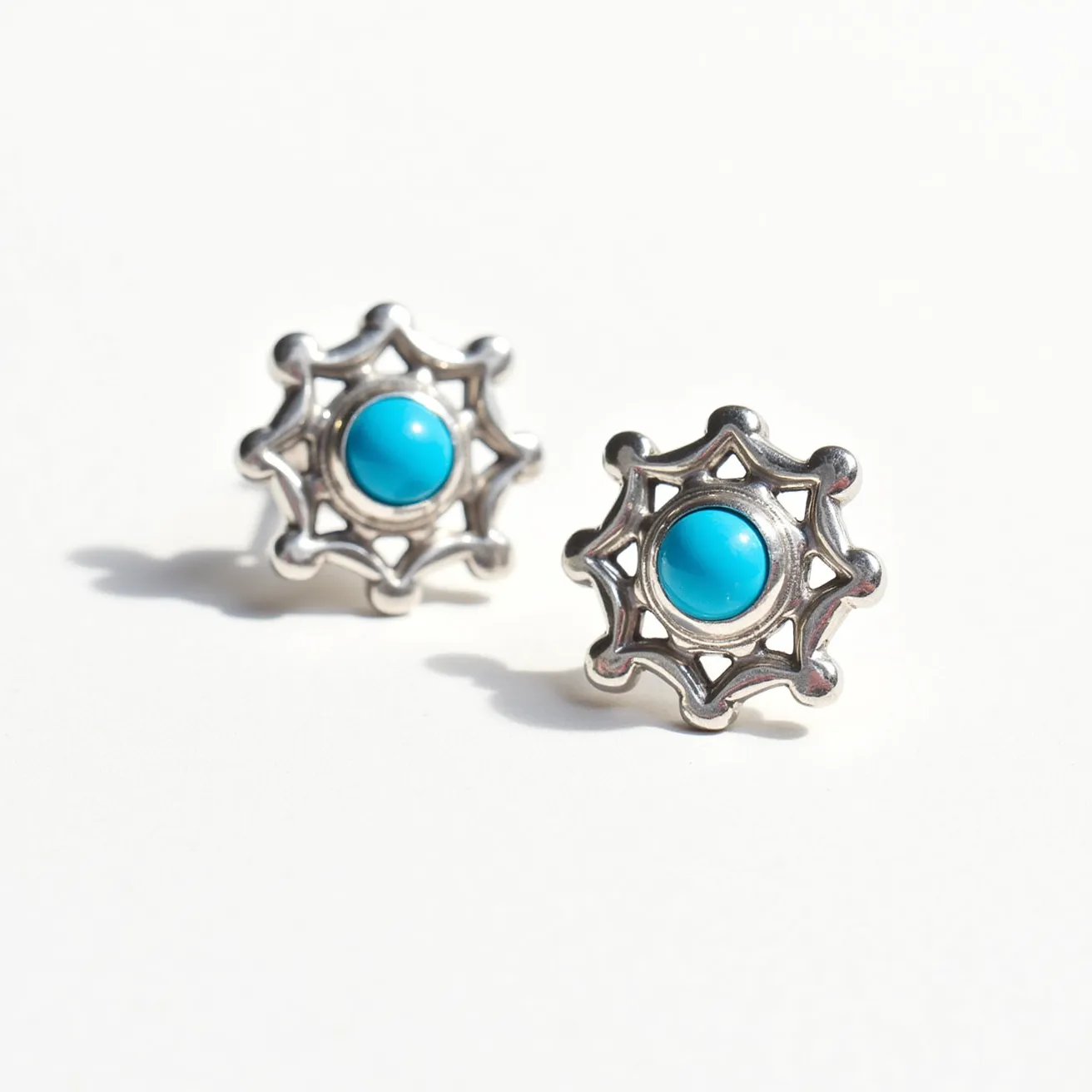 These turquoise stud earrings feature a central turquoise gemstone, which is smoothly polished and set in a bezel setting. The earrings are crafted from what appears to be a silver-toned metal, forming an intricate, openwork star-like design that surrounds the stone. The combination of the vibrant turquoise against the metallic setting creates a striking contrast. These earrings are designed as studs, with a traditional post and friction back attachment for secure wear. The overall design is both elegant and versatile, suitable for various occasions.