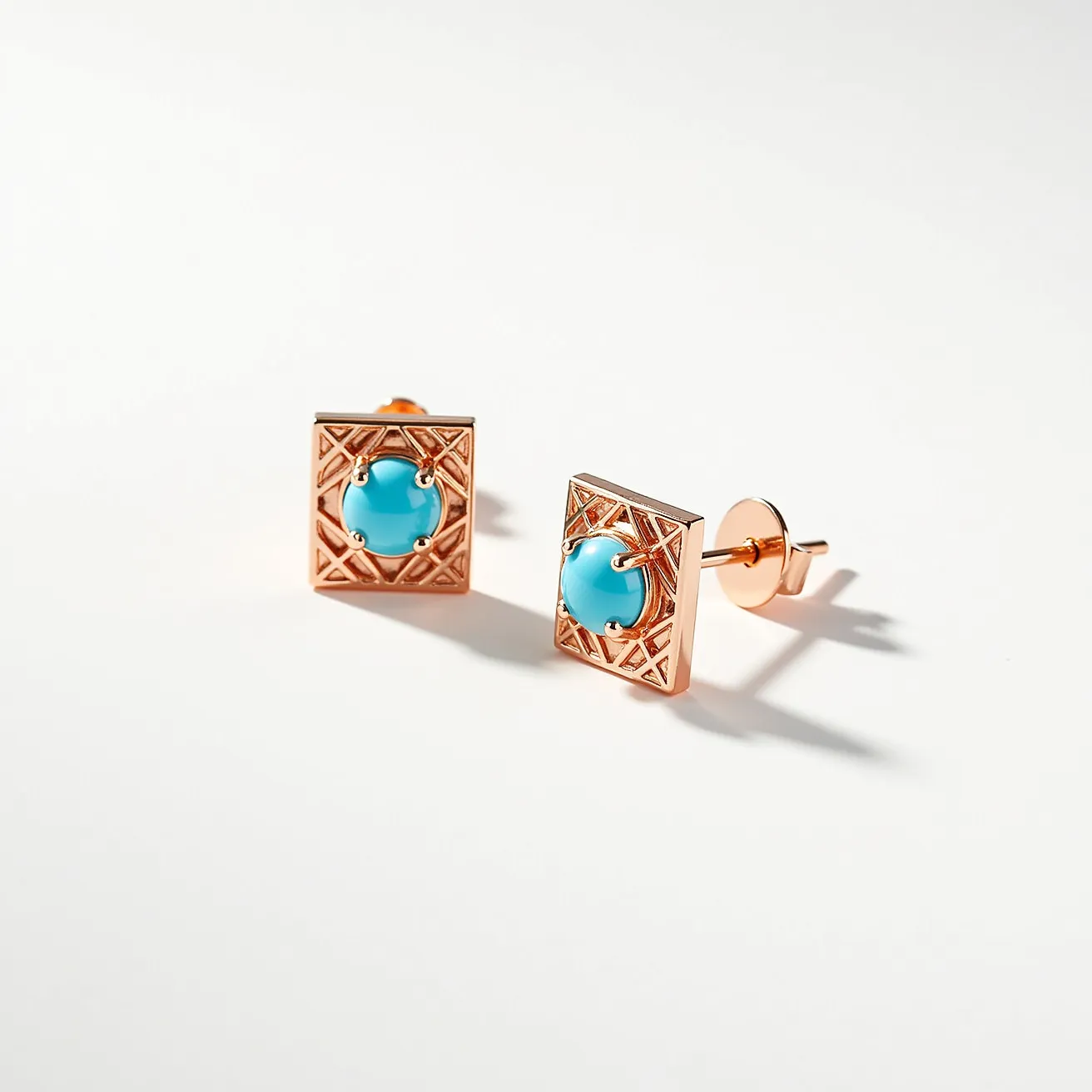 These turquoise stud earrings feature a striking turquoise gemstone, centrally set in a round cabochon cut. The stone is secured by a four-prong setting made of a rose-gold-toned metal that complements the vibrant blue hue of the turquoise. These earrings are adorned with a rectangular decorative frame that has an intricate geometric pattern, adding an elegant and artistic touch to the design. The studs are equipped with a classic post and friction back attachment, ensuring secure and comfortable wear.