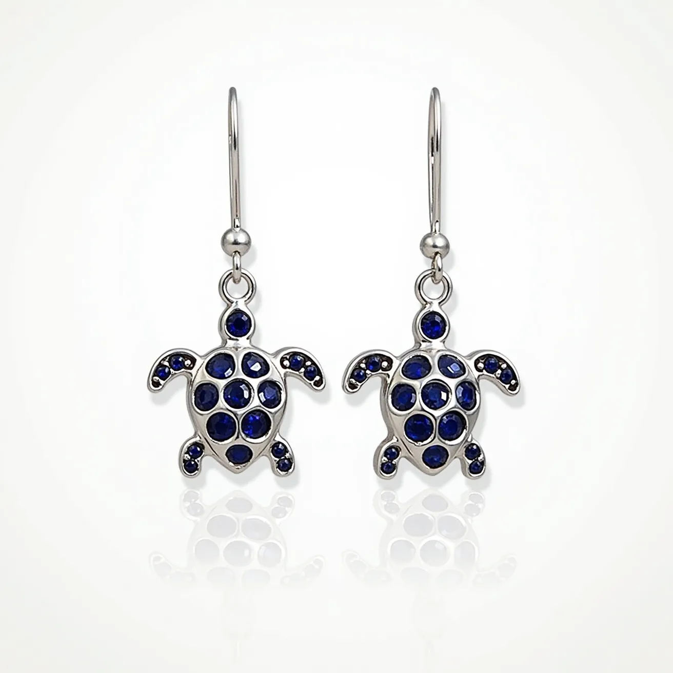 These turtle earrings feature a charming design where the body and flippers of the turtles are embedded with deep blue stones, likely sapphires, which are round-cut and set in a polished silver-toned metal. The stones are arranged to mimic the intricate design of a turtle's shell and limbs, adding a captivating contrast against the shimmering metal. These earrings utilize a simple hook attachment, which allows them to dangle freely and elegantly when worn. The combination of blue stones and silver-toned metal creates a harmonious, aquatic-themed aesthetic.