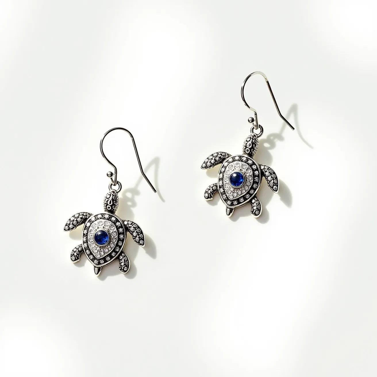 These turtle earrings feature a charming design crafted from a metallic material, likely silver, highlighting the intricate details of each turtle. The body of each turtle is adorned with a central blue gemstone, probably a sapphire, cut in a round shape and set in a bezel setting. Surrounding the blue stone are multiple small, clear stones that mimic diamonds, enhancing the overall sparkle. The earrings utilize a classic fishhook clasp for attachment, ensuring a secure and comfortable fit when worn.