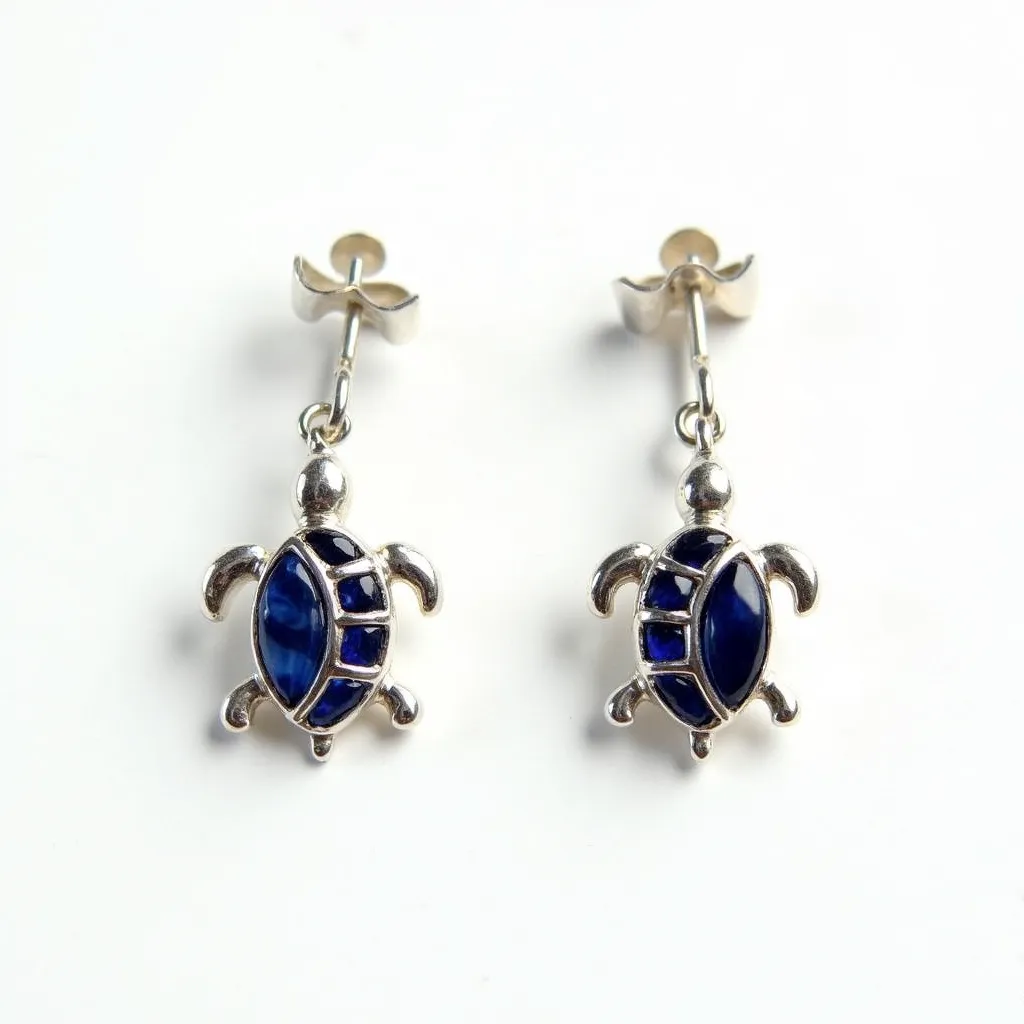 These turtle earrings feature a charming design with what appears to be a silver-toned metal crafting the shape of a turtle. The body of each turtle is adorned with blue gemstones, likely glass or synthetic stones, cut in a cabochon or smooth style, and set within the metal structure to mimic a turtle's shell pattern. These stones are set flush within the metal, providing a seamless and polished surface. The earrings attach with a simple post and butterfly back clasp, ensuring a secure and comfortable fit for pierced ears. The playful design combined with elegant materials makes these earrings a unique accessory.