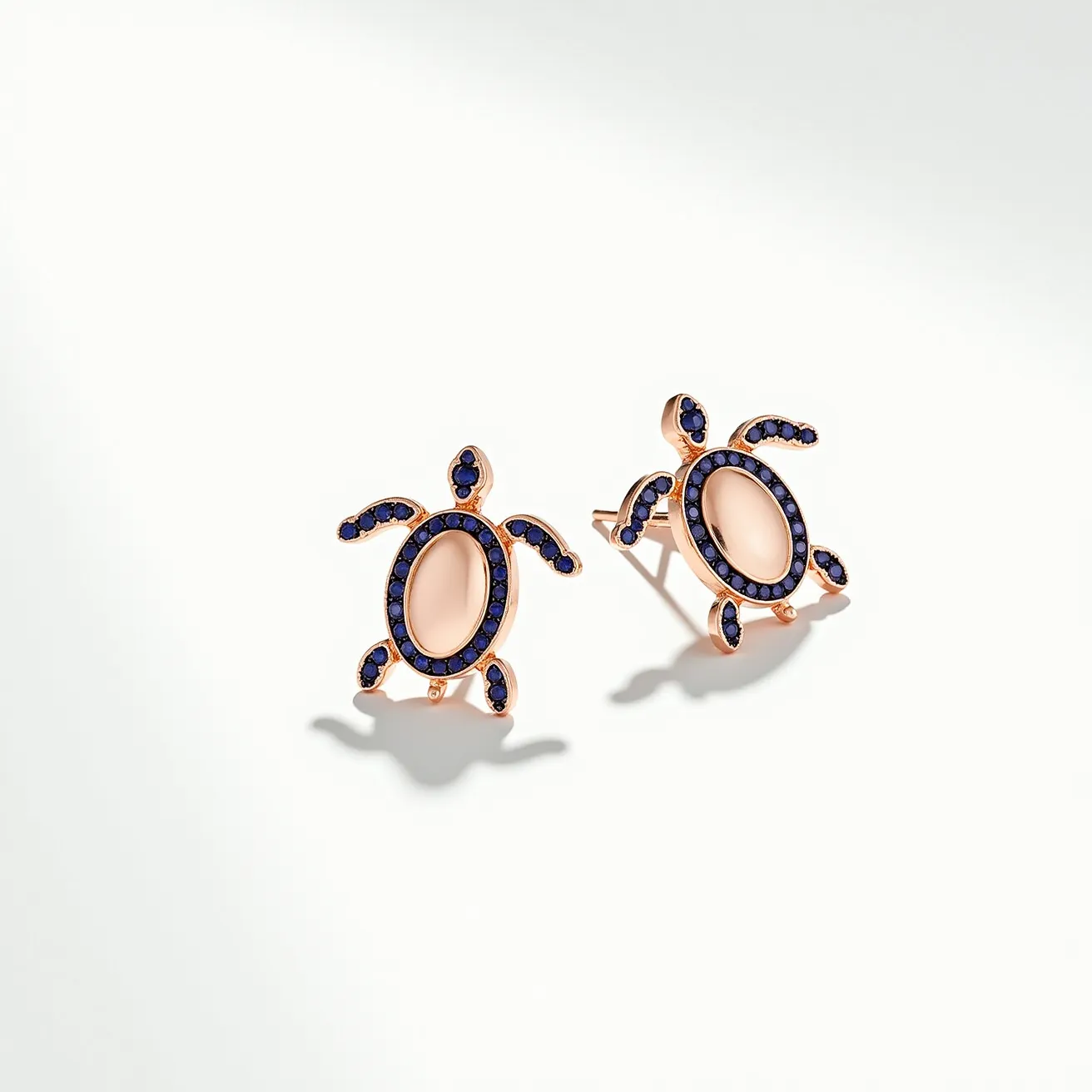 These turtle earrings feature a charming design with a rose gold-colored metal base forming the outline and structure of the turtle. The body of each turtle is accented with an oval-shaped, polished stone in a soft pink hue, adding a subtle elegance to the piece. Surrounding the body and adorning the flippers and head are small, round blue stones that bring a vibrant contrast, likely set using a pavé or micro setting technique. The earrings are designed with a simple post and butterfly clasp, ensuring a secure and comfortable fit when worn.