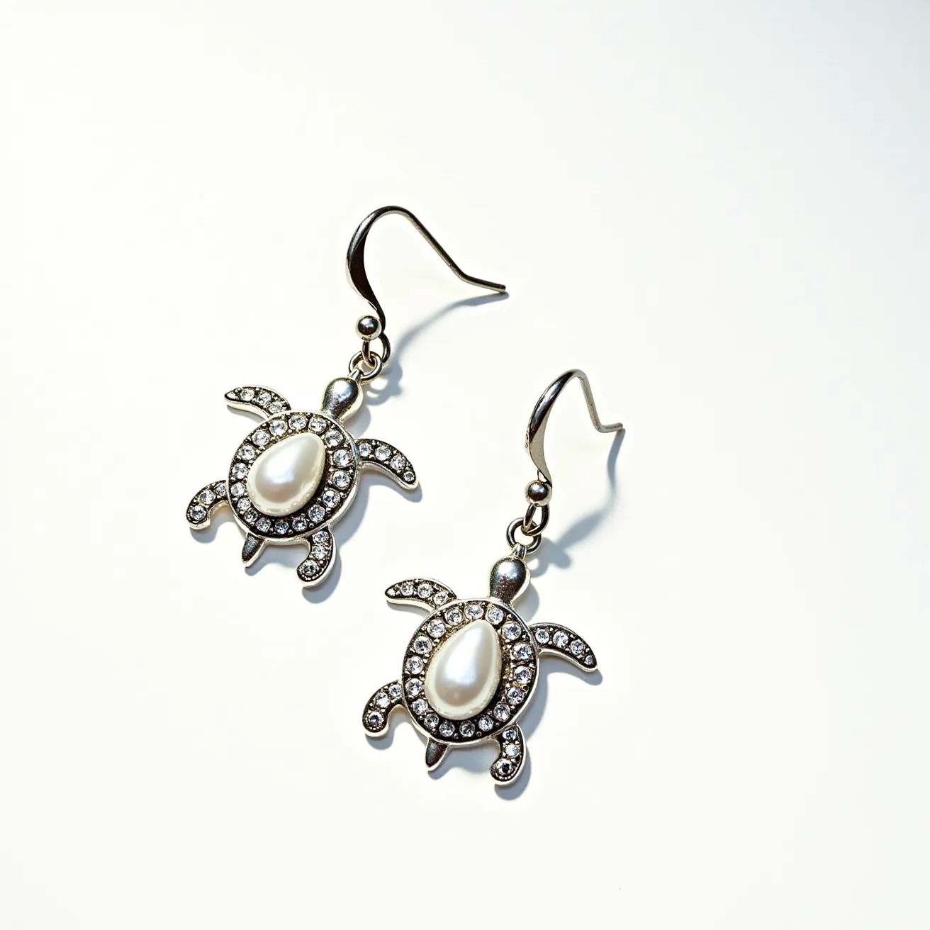 These turtle earrings are crafted with a metallic framework that outlines each turtle shape, featuring an elegant teardrop-shaped pearl at the center, embodying the turtle's shell. The surrounding metal is embellished with small, sparkling clear stones, likely crystals or diamonds, that are neatly set into the surface, enhancing the overall shine and luxury of the earrings. The earrings use a classic fishhook clasp that allows for easy attachment and comfortable wear. The detailed design and choice of materials combine to create a whimsical yet sophisticated accessory with a nod to nature.