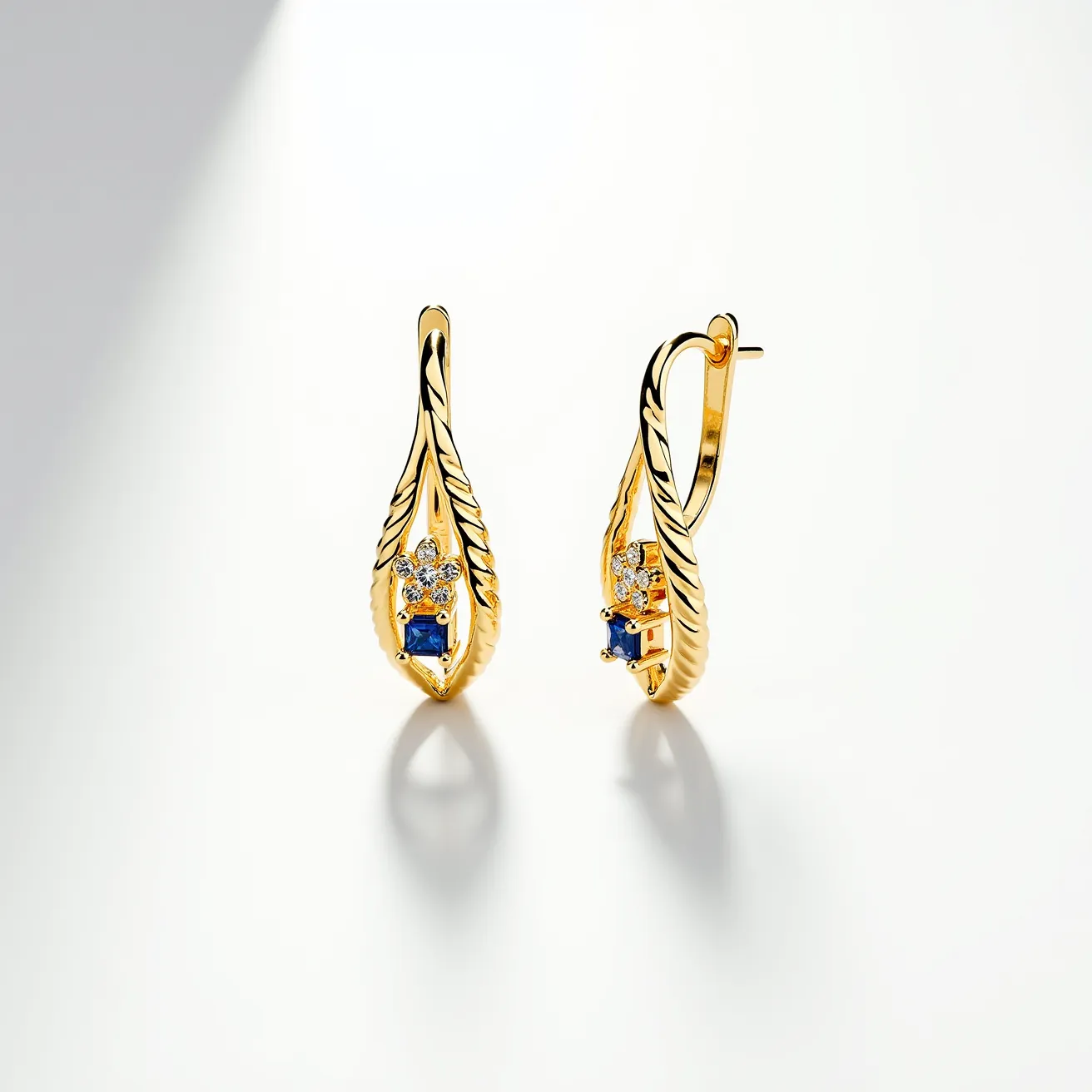 These twist earrings feature a graceful design crafted from gold, showcasing a twisted, leaf-like motif. At the core of each earring is a striking blue gemstone, likely a sapphire, cut in a square shape and securely set in a prong setting. Surrounding the central blue gemstone, there are several small, round clear stones that resemble diamonds, enhancing the overall elegance. The earrings are attached with a classic lever-back clasp, ensuring a secure and comfortable fit when worn. The intricate detailing along the gold surface adds to the luxurious appeal of these exquisite earrings.