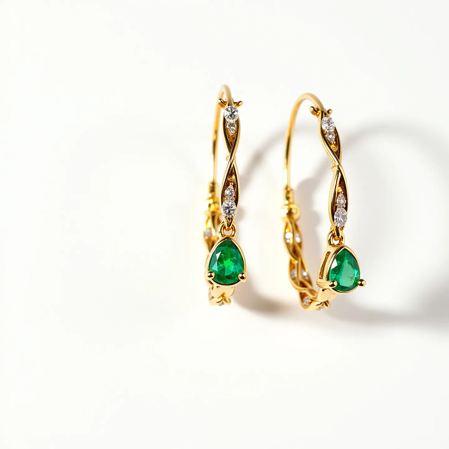 These twisted hoop earrings feature a sophisticated design crafted from gold, with an intricate twisting pattern along the hoop. Each earring is adorned with several small, brilliant-cut diamonds set into the gold, adding sparkle and elegance. At the bottom of each hoop hangs a pear-shaped emerald, held securely in a prong setting that highlights its vibrant green hue. The earrings are designed with a latch-back clasp, ensuring a secure and comfortable fit when worn. The combination of gold, diamonds, and emeralds gives these earrings an exquisite and luxurious appearance.