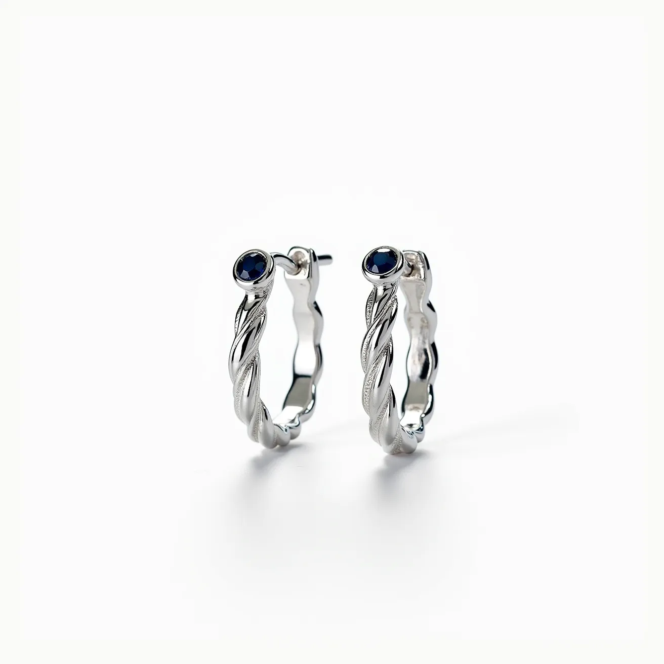 These twisted hoop earrings are crafted from a shiny metal, possibly silver, with a distinctive twisted design that adds texture and visual interest. Each hoop features a deep blue circular gemstone, likely a sapphire, set in a bezel setting, which provides a secure hold while highlighting the gem's color. The earrings are equipped with post backings, ensuring a snug and secure fit through the earlobe. The combination of the twisted metalwork and striking gemstones creates a sophisticated and elegant appearance.