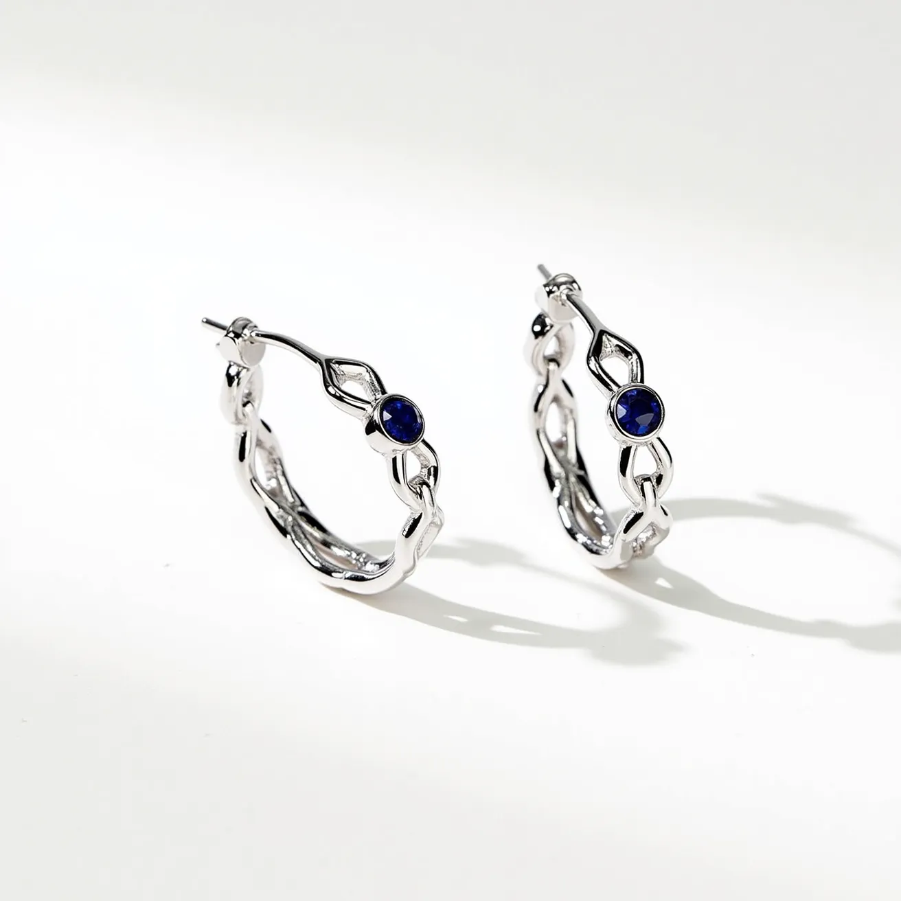 These twisted hoop earrings exhibit a sleek, intertwined design crafted from a shiny metal, possibly sterling silver or white gold, which enhances their elegant appearance. Each earring is adorned with a single deep blue gemstone, likely a sapphire or similar stone, set in a bezel setting that securely holds the stone while showcasing its vibrant color. The earrings feature a post and butterfly back clasp, ensuring a secure fit when worn. The intricate twisting and the gemstone placement add a sophisticated touch to the overall design.