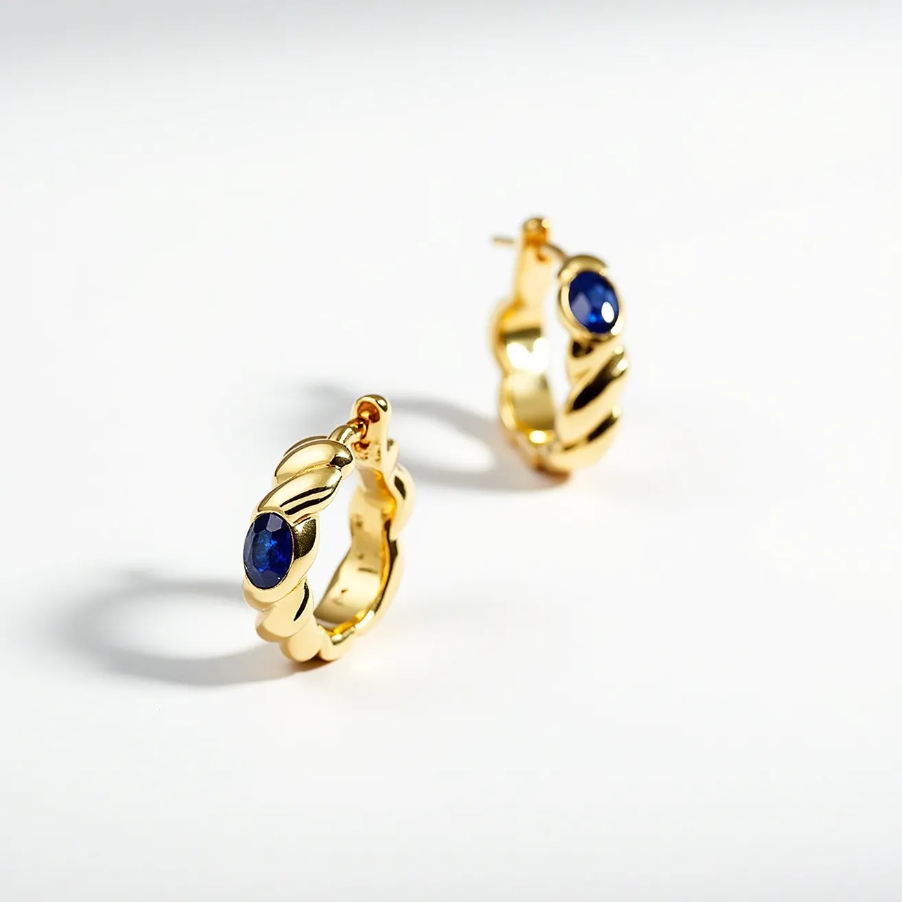 These twisted hoop earrings exhibit a luxurious and elegant design crafted from a polished gold material. The hoops are adorned with a deep blue gemstone, likely sapphire, that is set in a bezel setting, providing both security and a seamless look. The gemstones are cut in an oval shape, accentuating their rich color and clarity. The earrings feature a hinged clasp mechanism, ensuring ease of wear and secure fastening. The gold twists add a dynamic texture that highlights the sophistication and artistry of the design.