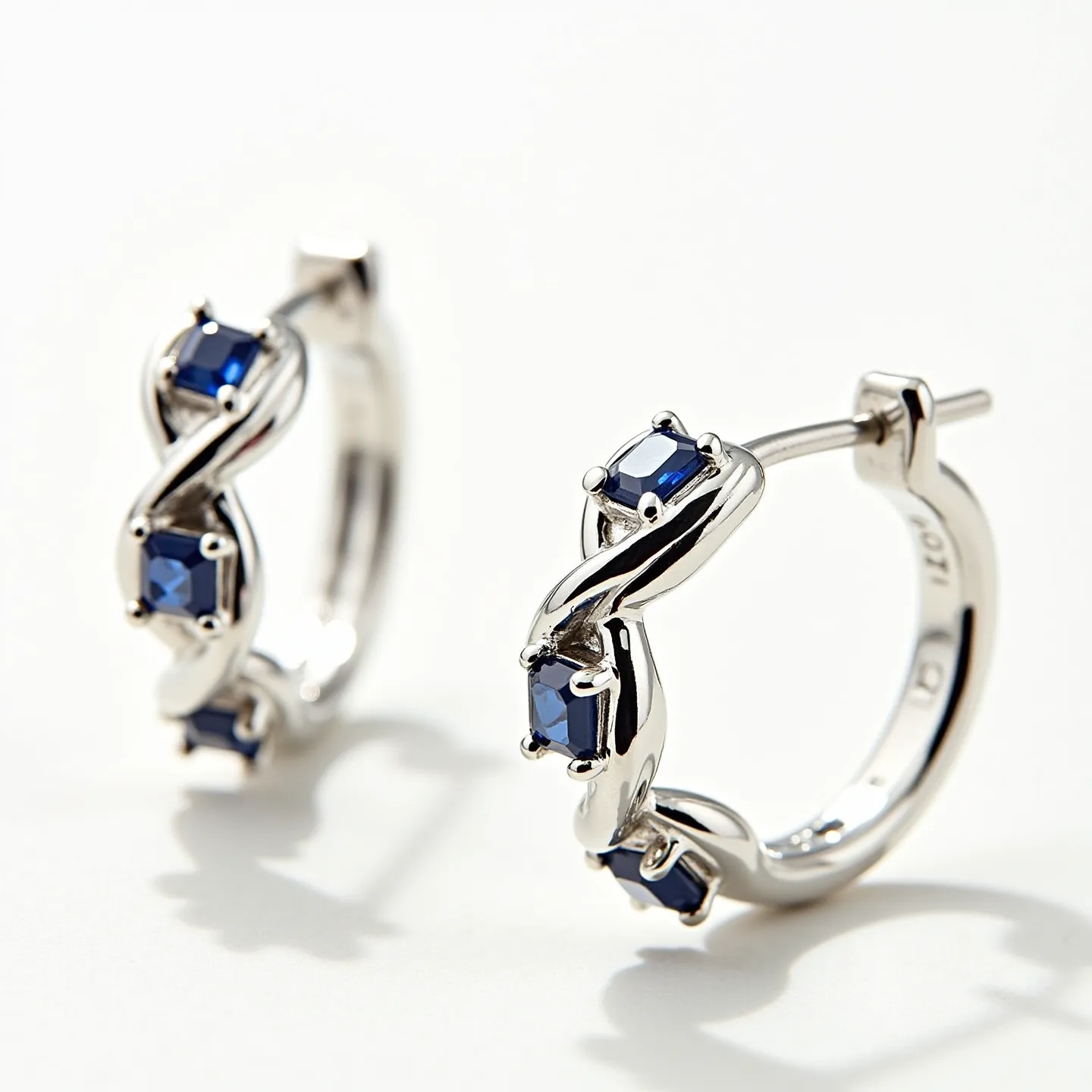 These twisted hoop earrings feature an elegant design crafted from silver-toned metal that encircles the ear in a smooth, flowing pattern. The earrings are adorned with square-cut blue gemstones securely set within prongs, adding a touch of color and sophistication to the piece. The hoops utilize a post and latch back clasp for secure fastening, ensuring comfort and ease of wear. The intricate twist of the metal adds a dynamic visual element, beautifully complementing the deep blue of the stones.