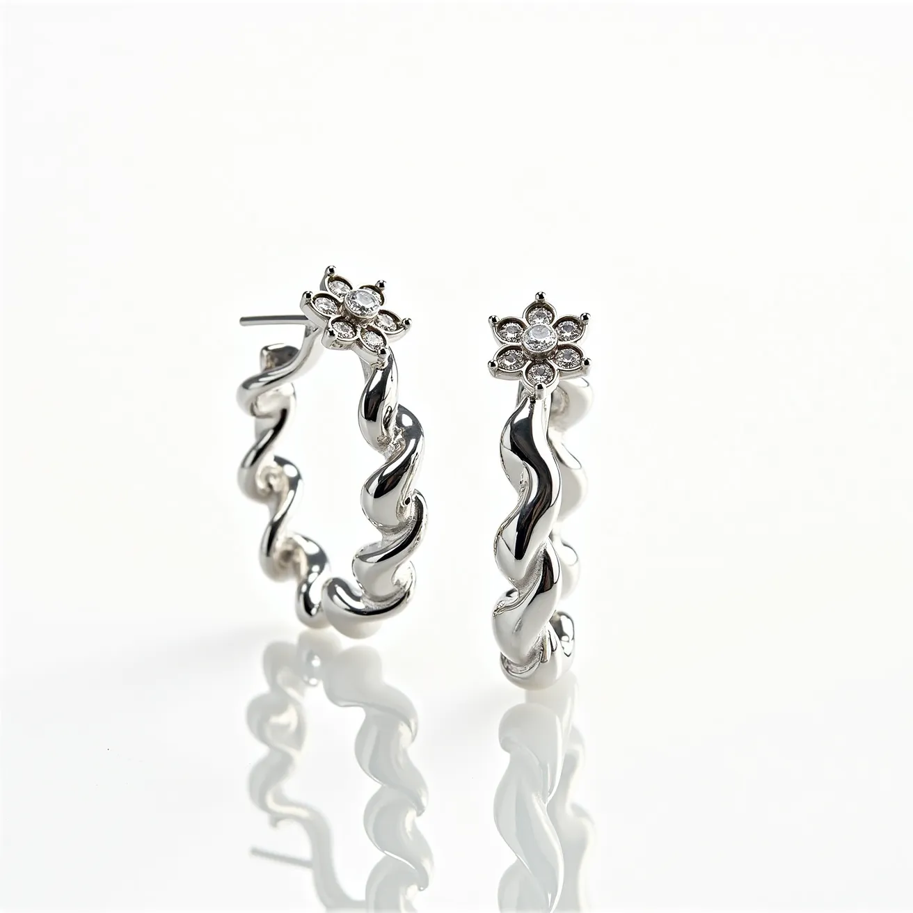 These twisted hoop earrings are crafted from a polished metal with a sleek, reflective surface, likely silver or white gold, giving them an elegant and contemporary look. The design features a distinctive twisted pattern that adds a dynamic texture to the hoops. Each earring is adorned with a flower-shaped cluster of small, round stones, likely diamonds, set in a prong setting at the top. The stones are brilliant cut, creating a sparkling focal point that enhances the overall design. The earrings are equipped with a straight post, suggesting a secure friction back or push-back clasp to ensure they stay in place when worn.