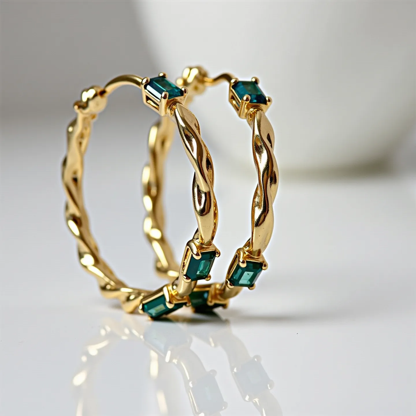 These twisted hoop earrings are crafted from a shiny gold material, featuring a distinctive twisted design that adds a dynamic element to their appearance. They are adorned with rectangular green gems set in a bezel setting that secures them along the hoops at regular intervals. The gems are precisely cut, enhancing their vibrant color and reflective qualities. The earrings are equipped with a simple hinge clasp that ensures secure and easy wear.