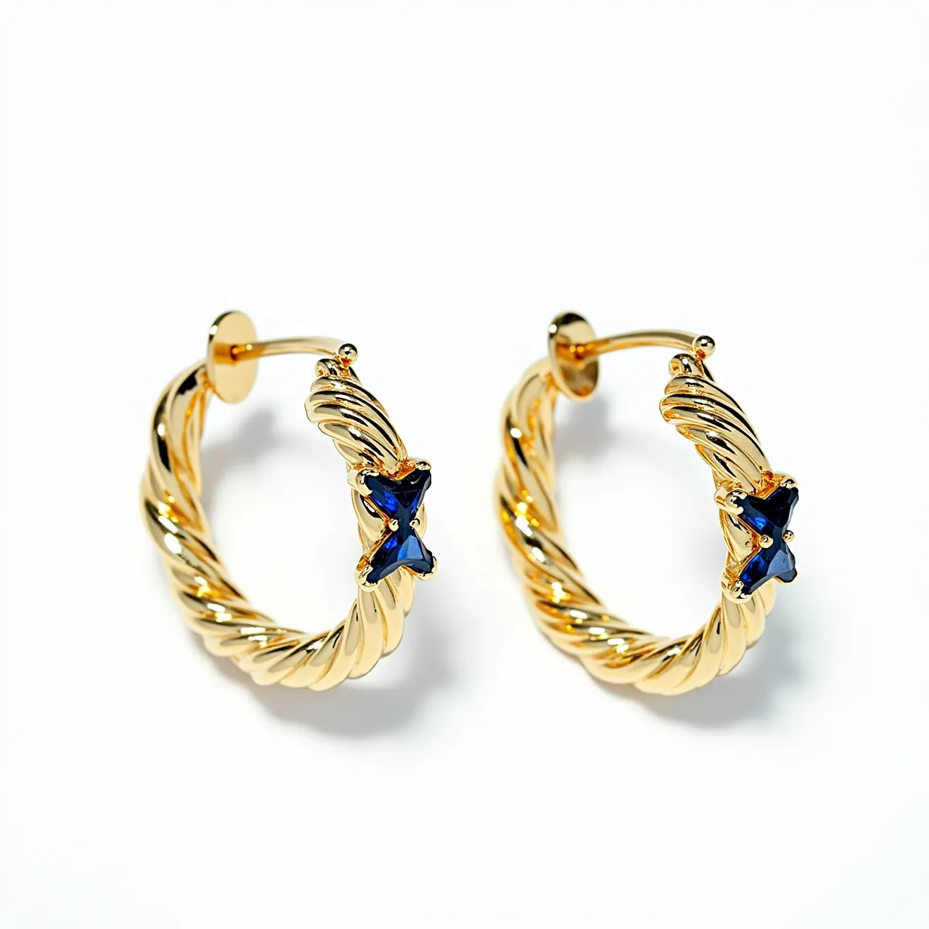 These twisted hoop earrings feature a design crafted from a shiny gold-toned material, forming an elegant twisted pattern. Each earring is adorned with a deep blue, X-shaped gemstone, likely a sapphire, set at the forefront for a striking contrast. The gemstones are secured in a prong setting, which enhances their visibility and brilliance. The earrings use a post and back clasp attachment, ensuring a secure fit while being worn.