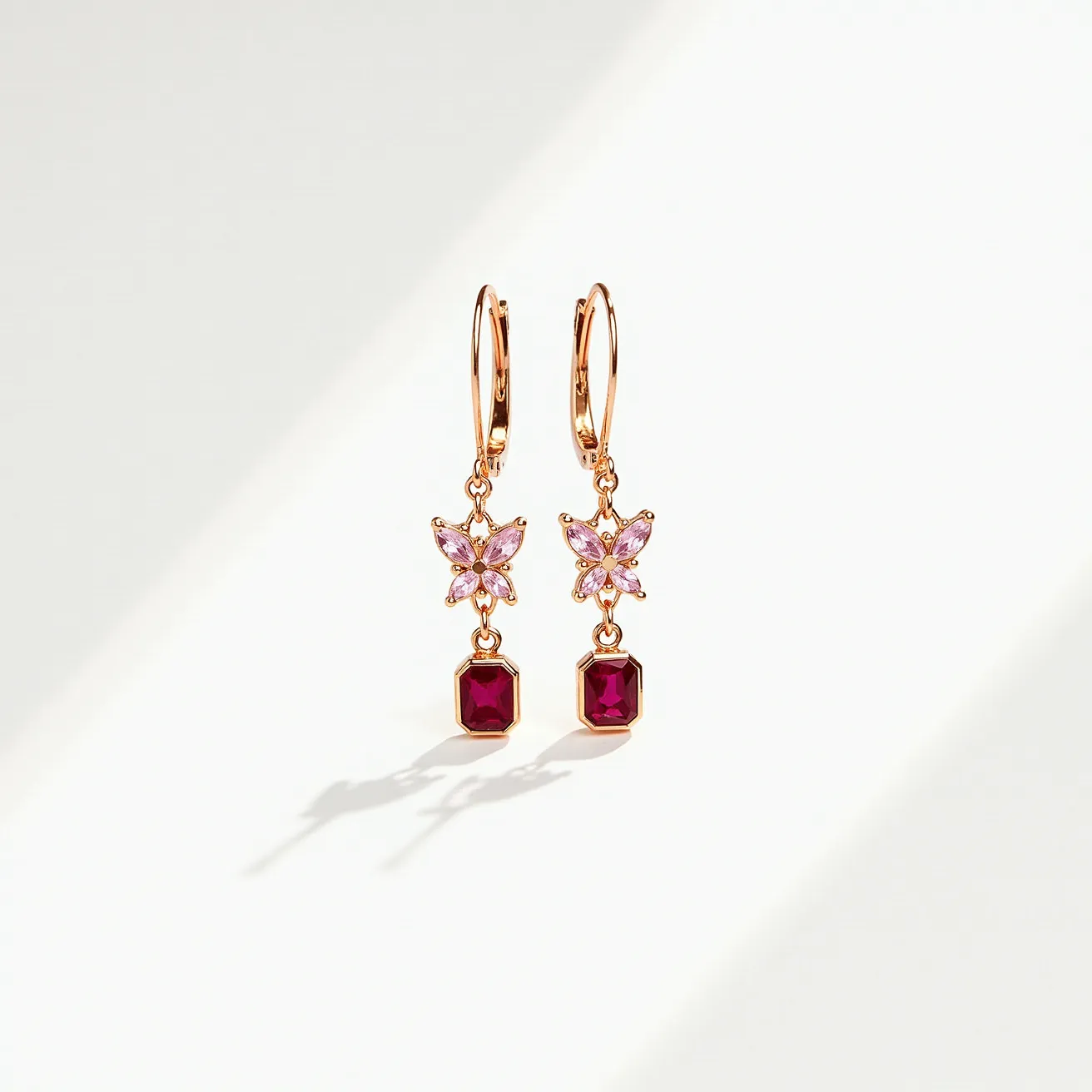 These valentines earrings feature a delicate design composed of rose gold-colored metal. They showcase butterfly shapes adorned with small pink gemstones, likely in a marquise cut, creating a floral effect. Hanging beneath each butterfly is a vibrant red gemstone, cut in an emerald shape, securely set in a bezel setting. The earrings are equipped with lever-back clasps, ensuring a secure and refined fastening.