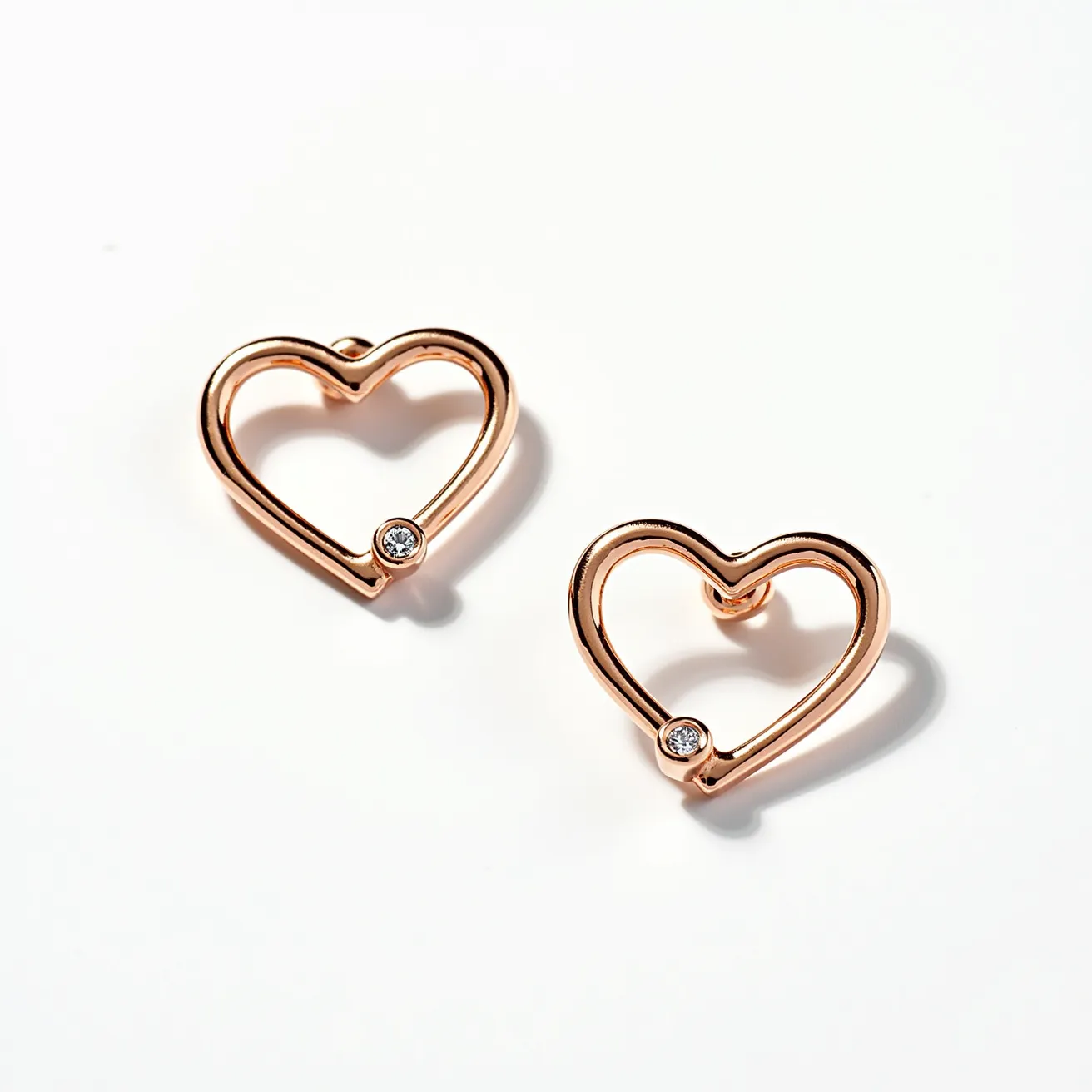 These valentines earrings feature a charming open-heart design crafted from a warm, rose gold metal. Each heart is embellished with a small, round white stone, likely a diamond or a cubic zirconia, set in a bezel setting situated at the bottom curve of the heart. The earrings are designed to be studs, with a post attachment visible from the back, suited for pierced ears and secured by a friction back clasp. The simplicity and elegance of the design make them a perfect accessory for a romantic occasion.