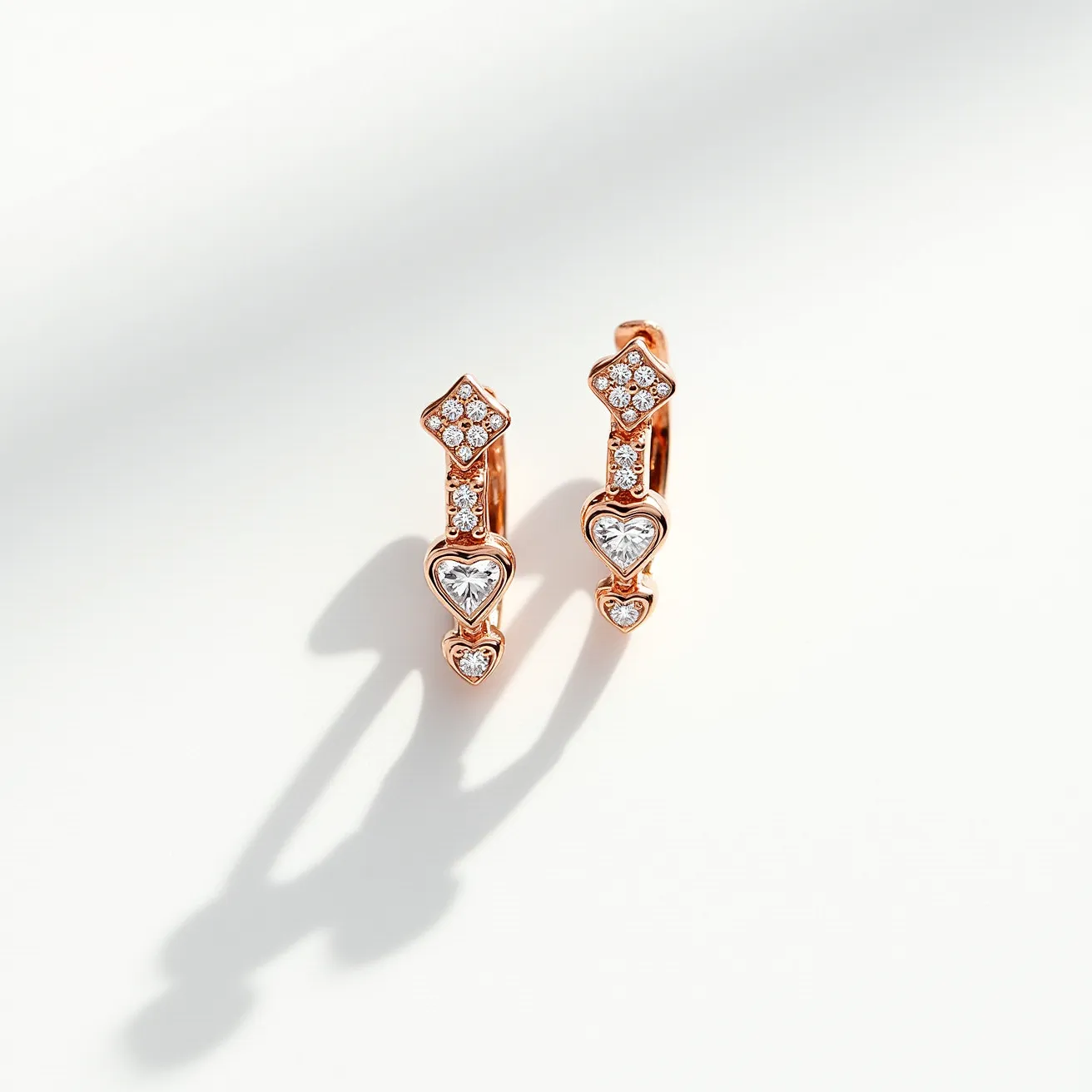 These valentines earrings features a romantic design crafted from a rose gold material, elegantly shaped into a series of geometric and heart motifs. Adorned with sparkling gems, these earrings showcase round cut diamonds in a pavé setting along the round diamond-shaped top. The focal point is a larger heart-shaped gem, also in a brilliant cut, securely set in the center, followed by a smaller heart-shaped gemstone at the bottom. The earrings employ a simple hoop attachment for secure and comfortable wear, highlighting both intricate craftsmanship and timeless elegance.

