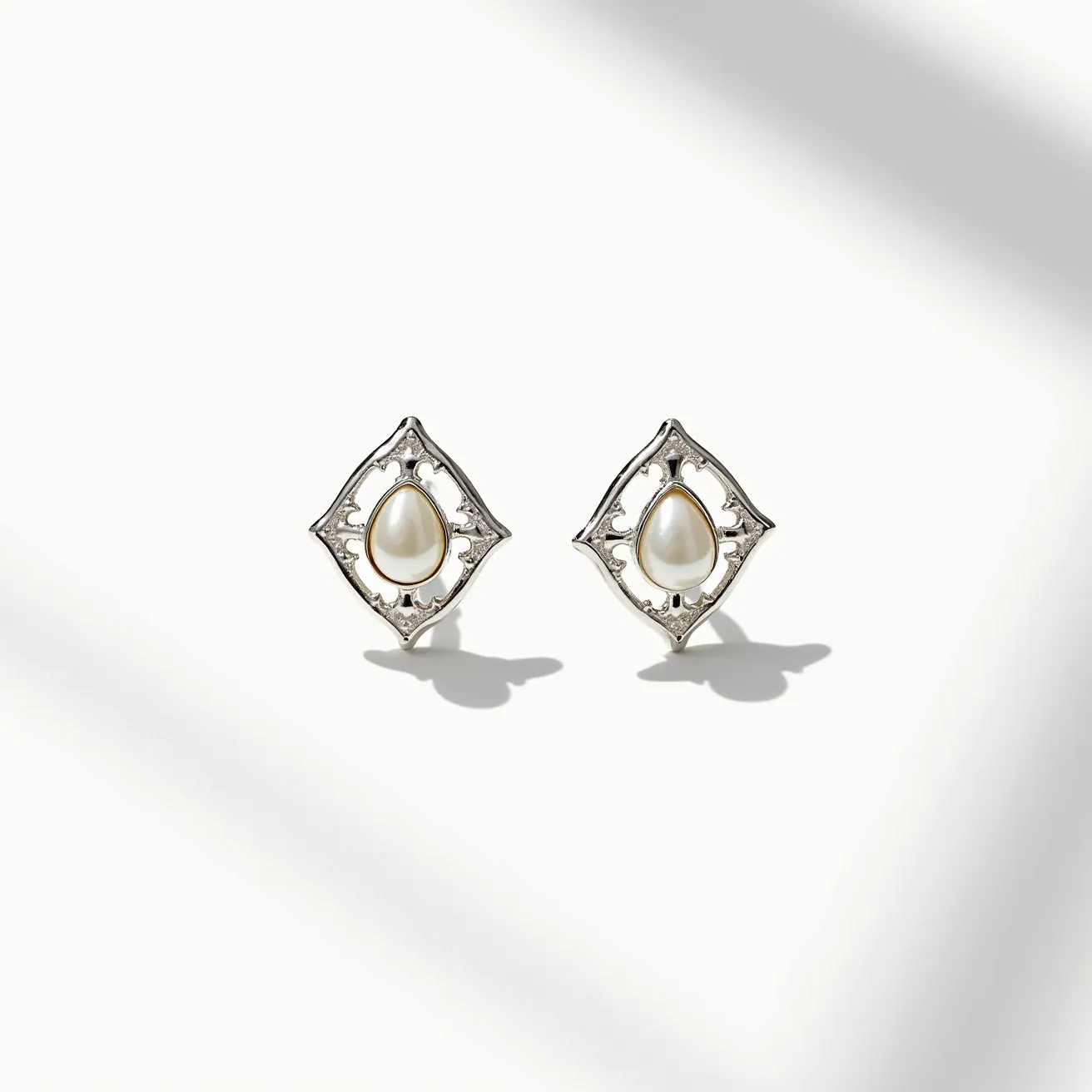 These white earrings feature an elegant design with a central white, teardrop-shaped gem that resembles a pearl. The gem is set in a polished metal frame that is intricately designed with lattice-like details, suggesting a classic and sophisticated style. The setting is open, allowing light to enhance the gem's lustrous quality. The earrings likely use a standard post or stud clasp for secure attachment, emphasizing both functionality and elegance. The overall design combines simplicity with intricate details, making them a versatile accessory for various occasions.