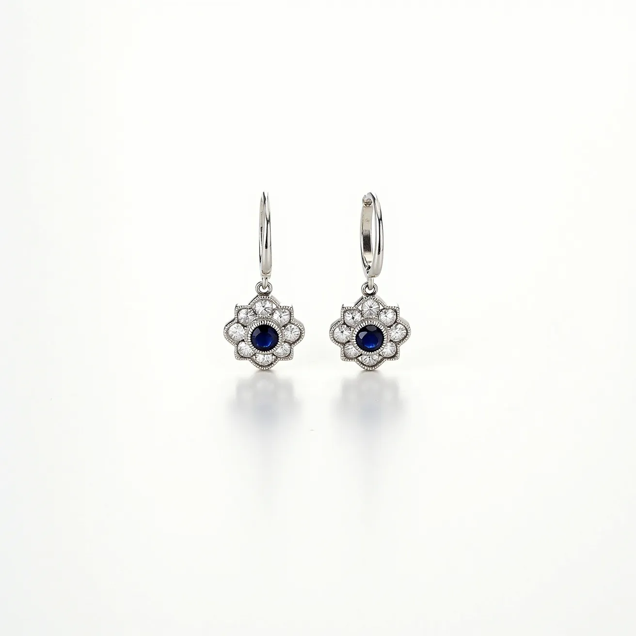 These white earrings feature an elegant floral design that incorporates a central blue sapphire, cut in a round style, and enclosed within a bezel setting. The surrounding petals of each earring are adorned with smaller white gemstones, which are neatly set to enhance the floral motif and add a brilliant contrast to the central stone. The earrings are crafted in a white metal, possibly white gold or platinum, which offers a lustrous shine. They include secure lever-back clasps, ensuring both comfort and safety when worn. These earrings combine timeless design with a sophisticated touch, making them suitable for both everyday wear and special occasions.