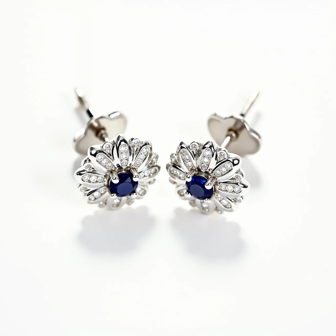 These white earrings feature a captivating design with a central deep blue gemstone, likely a sapphire, set in a prong setting that highlights its round cut. Surrounding the central stone are numerous small, clear stones, possibly diamonds, which are also set in prongs, creating a floral pattern that enhances the earrings' elegance. The earrings appear to be crafted from a shiny metal, possibly white gold or platinum, which complements the brilliance of the stones. The attachment mechanism is a classic stud with a butterfly clasp, ensuring secure and comfortable wear.