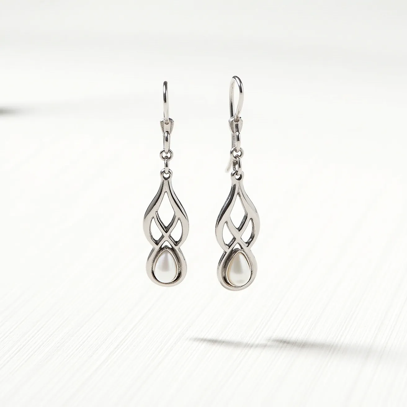 These white earrings feature a sleek and elegant design crafted from polished metal, possibly silver or white gold. Each earring showcases an openwork teardrop-shaped frame with intricate detailing, culminating in a single, oval-cut white pearl or stone securely set at the bottom of the design. The earrings are suspended from a simple hook clasp, providing ease of wear and allowing them to dangle freely, enhancing their graceful movement and sophisticated appeal.