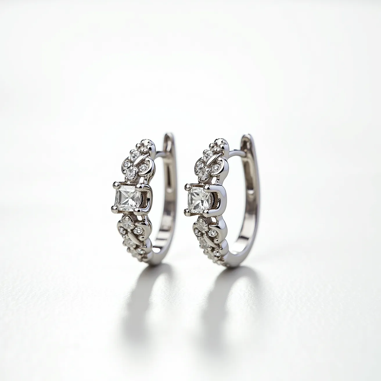 These white gold earrings feature a stunning design accentuated by a series of brilliant-cut diamonds. Each earring highlights a central princess-cut diamond, held in a secure prong setting that ensures the stone's stability while maximizing its sparkle. Surrounding the central stone, smaller round diamonds are meticulously set along the curve of the earring, adding an elegant shimmer. The earrings are designed with a hinged hoop clasp for secure attachment, combining both functionality and style in a compact form.