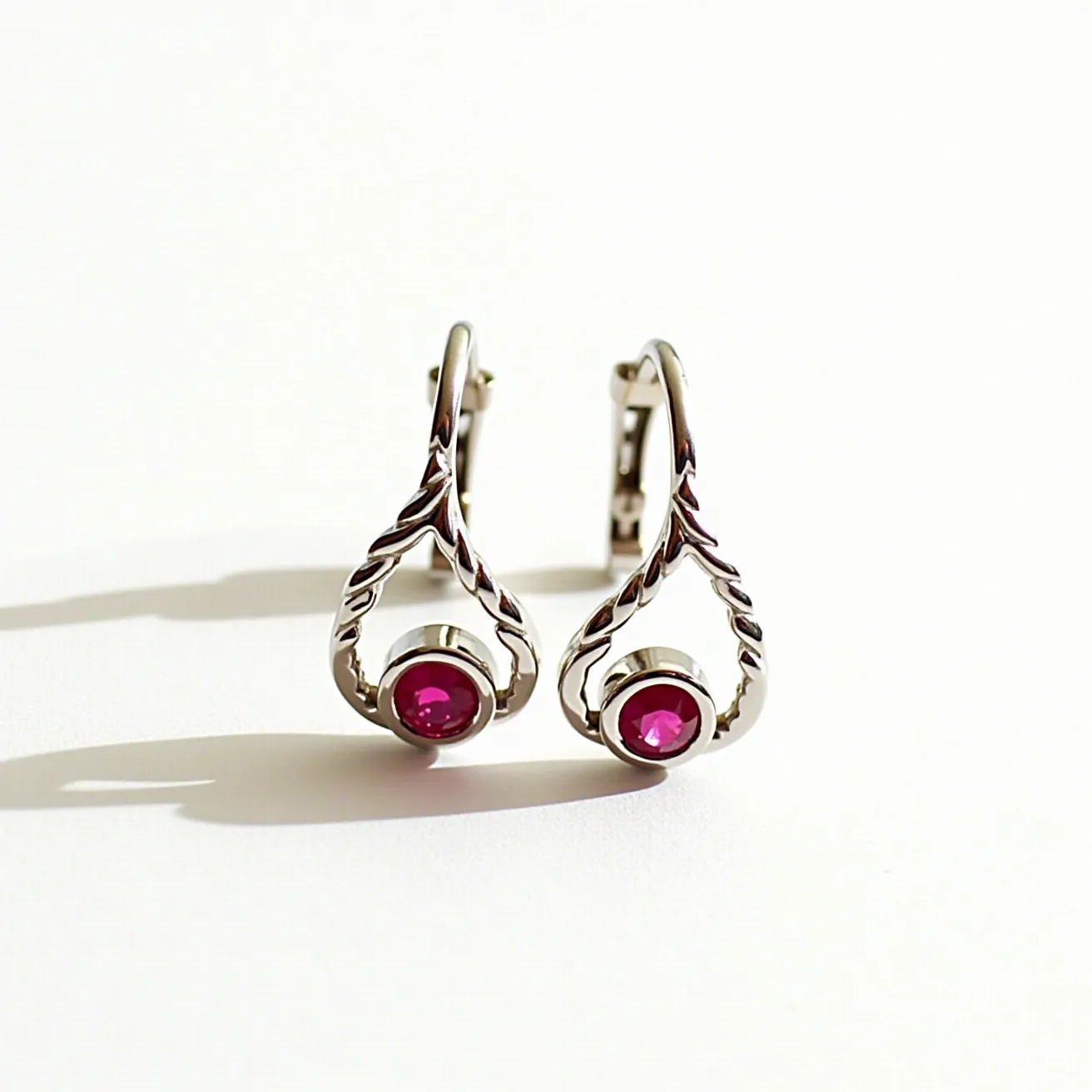 These white gold earrings feature a delicate and intricate design with a leaf-like motif elegantly curving around each side. At the center of each earring is a round-cut, deep red gemstone, likely a ruby, beautifully set in a bezel setting that enhances its vibrant color. The ornate leafy pattern adds a touch of elegance and sophistication to the overall aesthetics. The earrings are secured with a hinged lever-back clasp, providing both security and comfort for the wearer. The combination of white gold and the striking red gemstones creates a harmonious and stylish accessory.