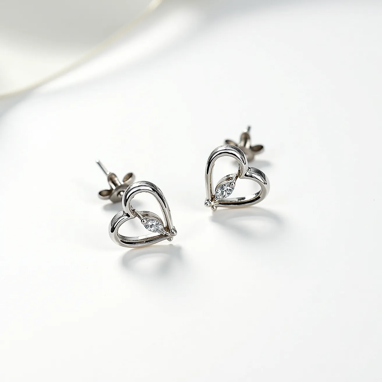 These white gold earrings feature an elegant heart design with each earring showcasing a single marquise-cut gemstone securely set at the center. The smooth, reflective surface of the white gold complements the brilliance of the gems, enhancing their sparkle. The earrings utilize a post and butterfly clasp attachment, ensuring a secure fit when worn. The overall design is delicate and sophisticated, making these earrings a versatile accessory suitable for various occasions.