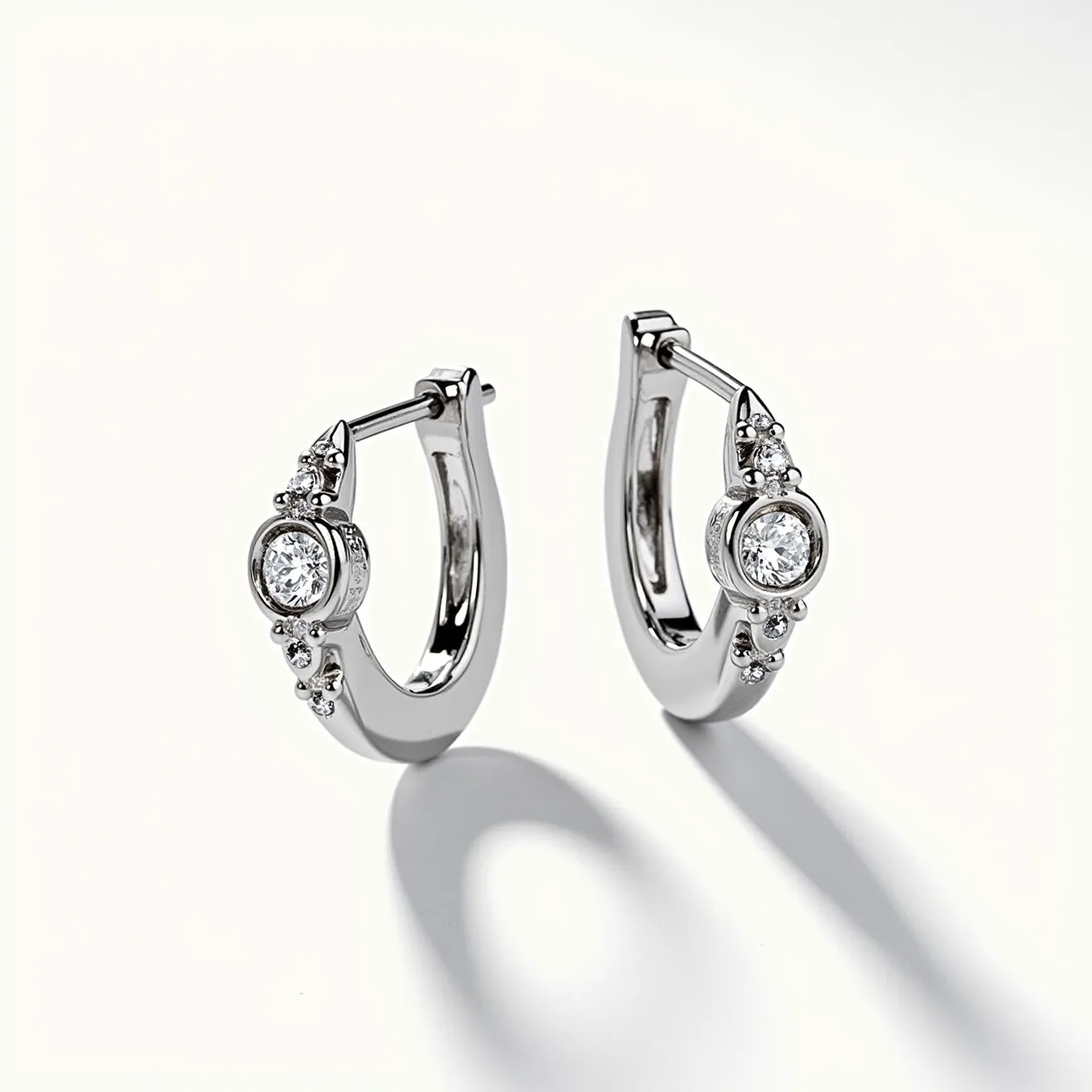 These white gold earrings feature a sleek, contemporary design with a prominent round brilliant-cut diamond securely set in a bezel setting. The central stone is complemented by smaller, round diamonds encircling it, creating a radiant, eye-catching effect. The earrings have a hinged hoop design with secure clasps that offer both elegance and practicality. The polished white gold enhances the sparkle of the diamonds, making these earrings a sophisticated accessory for any occasion.