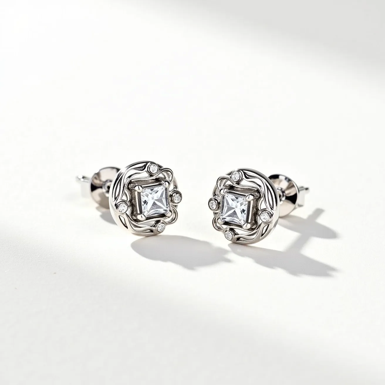 These white gold earrings feature a central square-cut gem, likely a diamond or a high-quality crystal, set in a sophisticated bezel setting. Surrounding the primary stone, a swirl pattern of white gold adds an elegant texture, further accentuated by smaller, round brilliant-cut stones nestled within the design. These decorative elements enhance the overall sparkle and elegance of the piece. The earrings are secured with a classic post and nut clasp, ensuring a secure fit while maintaining a sleek and modern aesthetic.