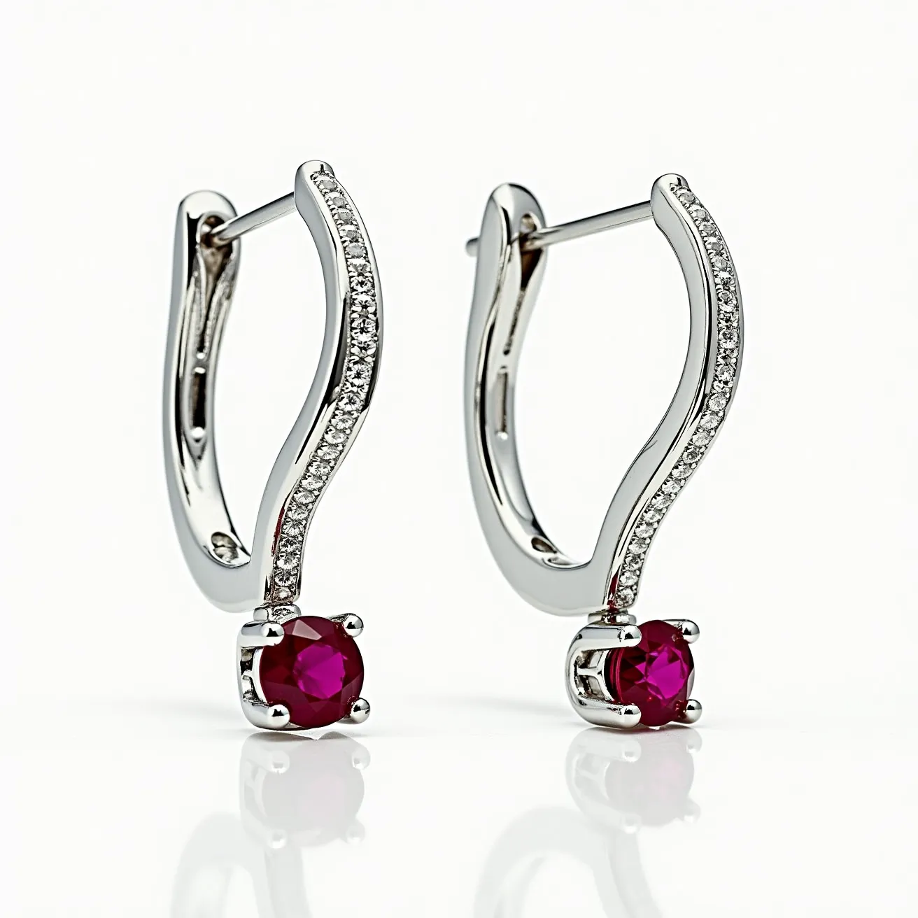 These white gold earrings feature an elegant design with a wave motif encrusted with small, round diamonds, adding a touch of sparkle to the overall aesthetic. At each tip of the earring, a round-cut ruby is set in a four-prong setting, providing a striking contrast with its deep red hue. These earrings utilize a hinged hoop clasp that ensures secure fastening while maintaining fluidity in design. The combination of the precious metals and gemstones creates a harmonious and luxurious piece of jewelry.