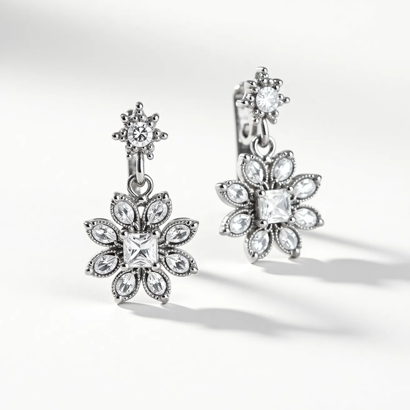 These white sapphire earrings feature an intricate design with a central square-cut white sapphire surrounded by six marquise-cut stones, mimicking a floral pattern. Each earring has a brilliant round-cut sapphire at the top, enhancing the sparkle and depth of the overall design. The stones are meticulously set in a polished silver metal that complements the clarity and brilliance of the sapphires. The attachment is a clip-on clasp, ensuring secure wear without the need for pierced ears. These elements come together to create an elegant and sophisticated accessory suitable for a variety of occasions.