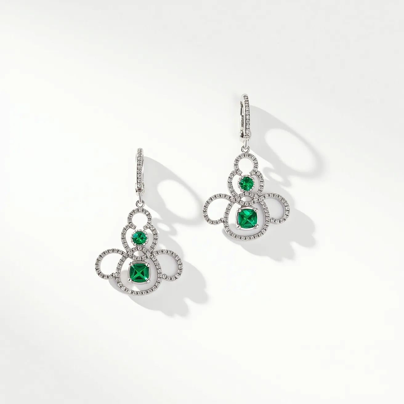 These wire earrings feature a delicate design crafted from a lustrous metal, possibly silver or white gold, creating an elegant looped pattern adorned with small, brilliant-cut gemstones that resemble diamonds. At the center of each looped section, there are vibrant green stones, likely emeralds, cut in a round and cushion shape, adding a pop of color and contrast against the metallic sheen. The earrings are designed with a hinged lever-back clasp, providing a secure yet comfortable fastening for the wearer. The overall composition exudes sophistication and intricate craftsmanship, making these earrings a striking accessory.