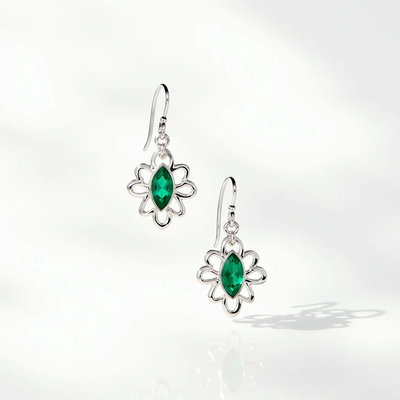 These wire earrings feature a delicate floral design crafted from a polished metal, likely silver. At the center of each earring, there is a prominent marquise-cut green gemstone, possibly an emerald or a similarly colored crystal, set in a bezel setting that securely holds the stone. The earrings have a fishhook attachment, offering ease of wear and a touch of elegance. The overall design combines a sparkling central gem with an intricate lace-like metal pattern, creating a balance of sophistication and graceful style.