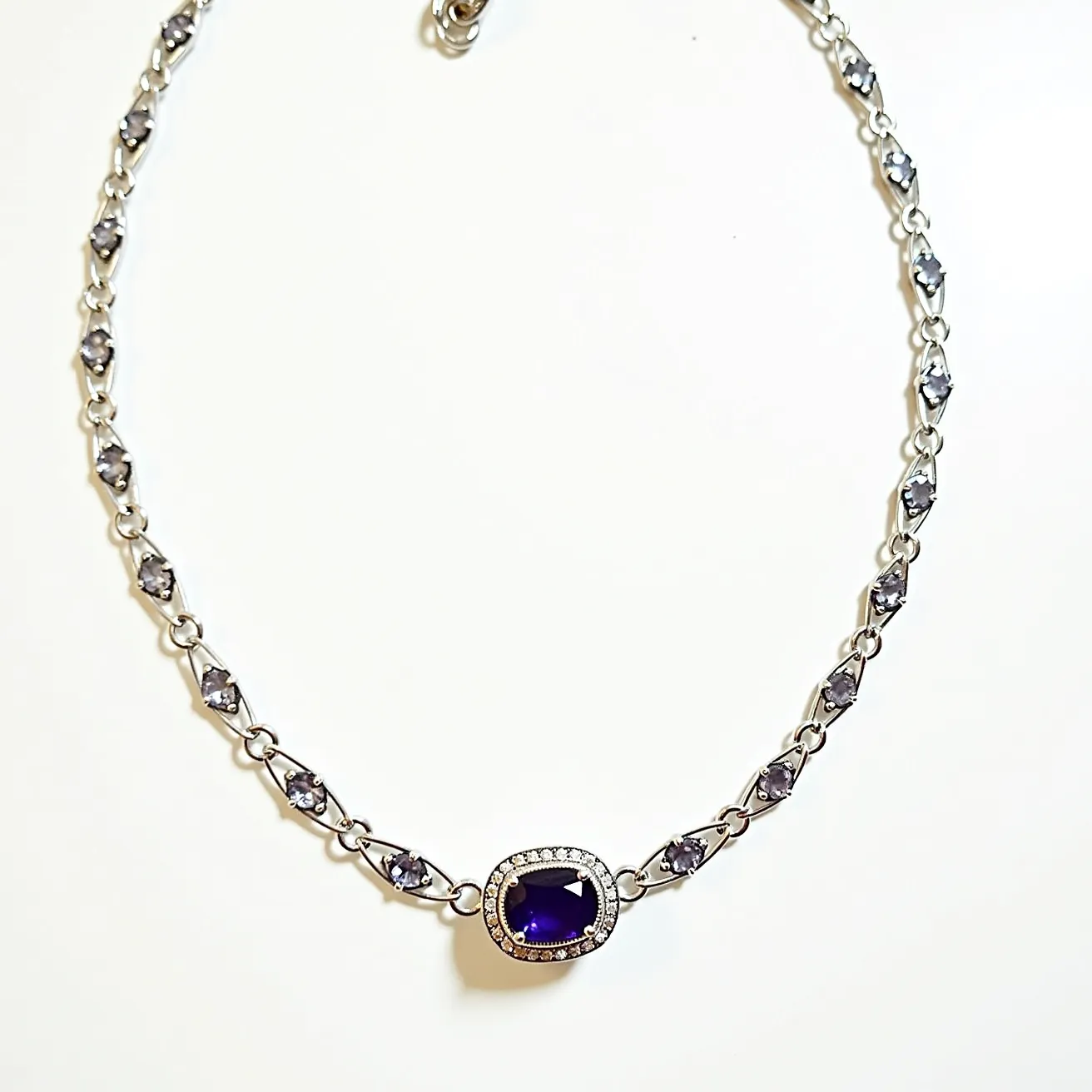 This alexandrite necklace features a striking central stone, possibly alexandrite, with a deep blue-violet hue set in a cushion cut. The stone is surrounded by a halo of smaller clear stones, enhancing its brilliance. The metal components appear to be made of a polished silver or white gold material, contributing to the necklace's elegant look. The chain consists of a series of similarly colored stones set in an alternating pattern within delicate links, adding to the overall allure. The piece is finished with a classic lobster clasp, ensuring secure wearability.