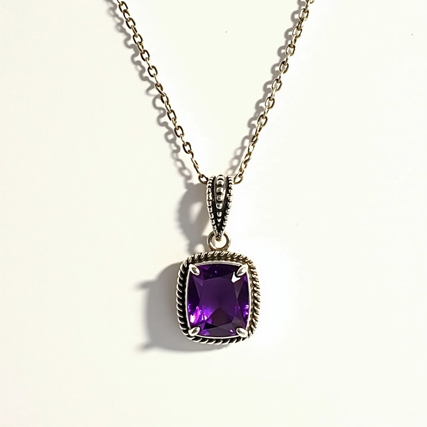 This amethyst necklace features a prominent rectangular amethyst stone, exhibiting a rich purple hue. The stone is cut in a cushion shape and is securely held in a prong setting. The pendant is adorned with a detailed metal border that includes an intricate rope-like design, enhancing the overall elegance of the piece. The necklace is attached to a delicate chain with a sturdy-looking clasp, and the bail connecting the chain to the pendant exhibits matching decorative features, adding to the necklace's cohesive and stylish appearance.