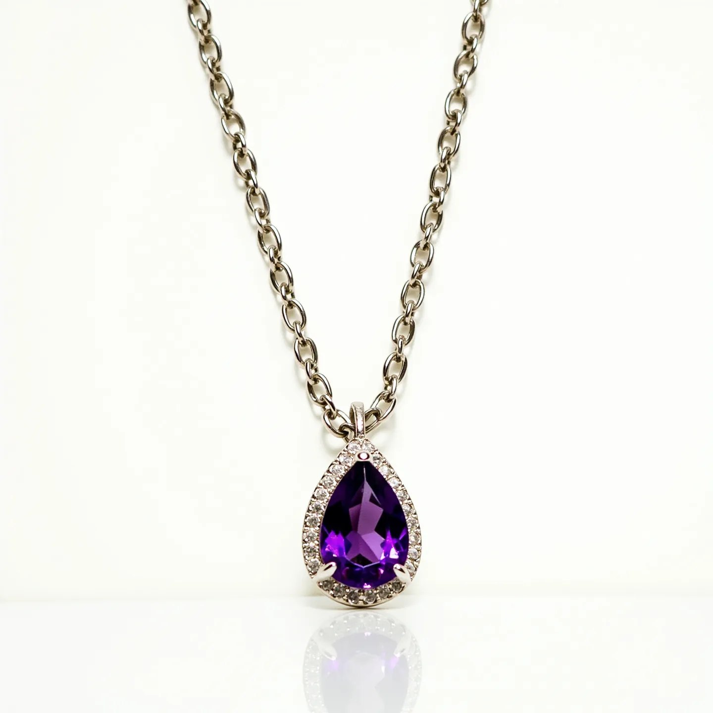 This amethyst necklace features a prominent pear-shaped amethyst gemstone as its centerpiece, showcasing a deep purple hue. The amethyst is elegantly set in a halo setting adorned with small, clear round stones, possibly diamonds or cubic zirconia, enhancing its brilliance. The pendant hangs from a robust chain, likely made of silver or white gold, which complements the cool tones of the stones. The chain appears to incorporate a secure, classic link structure, providing both strength and style to the piece. Overall, the combination of materials and design elements result in a sophisticated and eye-catching necklace.