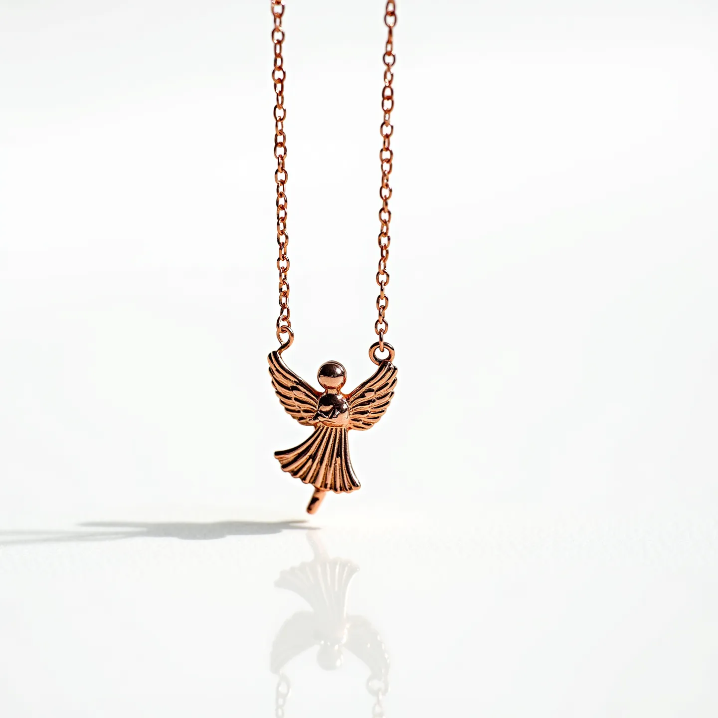 This angel necklace features a delicate chain made of a rose gold-toned metal, lending a warm and elegant appearance. The pendant is crafted in the shape of an angel, with detailed wings and a textured skirt design, also in the same rose gold tone. The angel's head is represented by a small, spherical bead, seamlessly integrated into the pendant's design. The pendant is attached to the chain via two small loops, one positioned at each wing tip. The necklace likely includes a standard clasp, though it's not visible in the image, ensuring secure wearability. The overall design combines simplicity and charm, making it a graceful accessory.